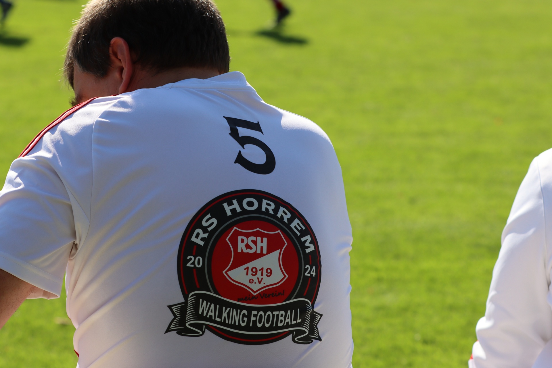 [{"clang_id":1,"value":"Rückansicht eines Fußballspielers mit weißem Trikot, Nummer 5 und Vereinslogo \"RSH Horrem 1924 Walking Football\" auf dem Rücken, stehend auf grünem Rasen."}]