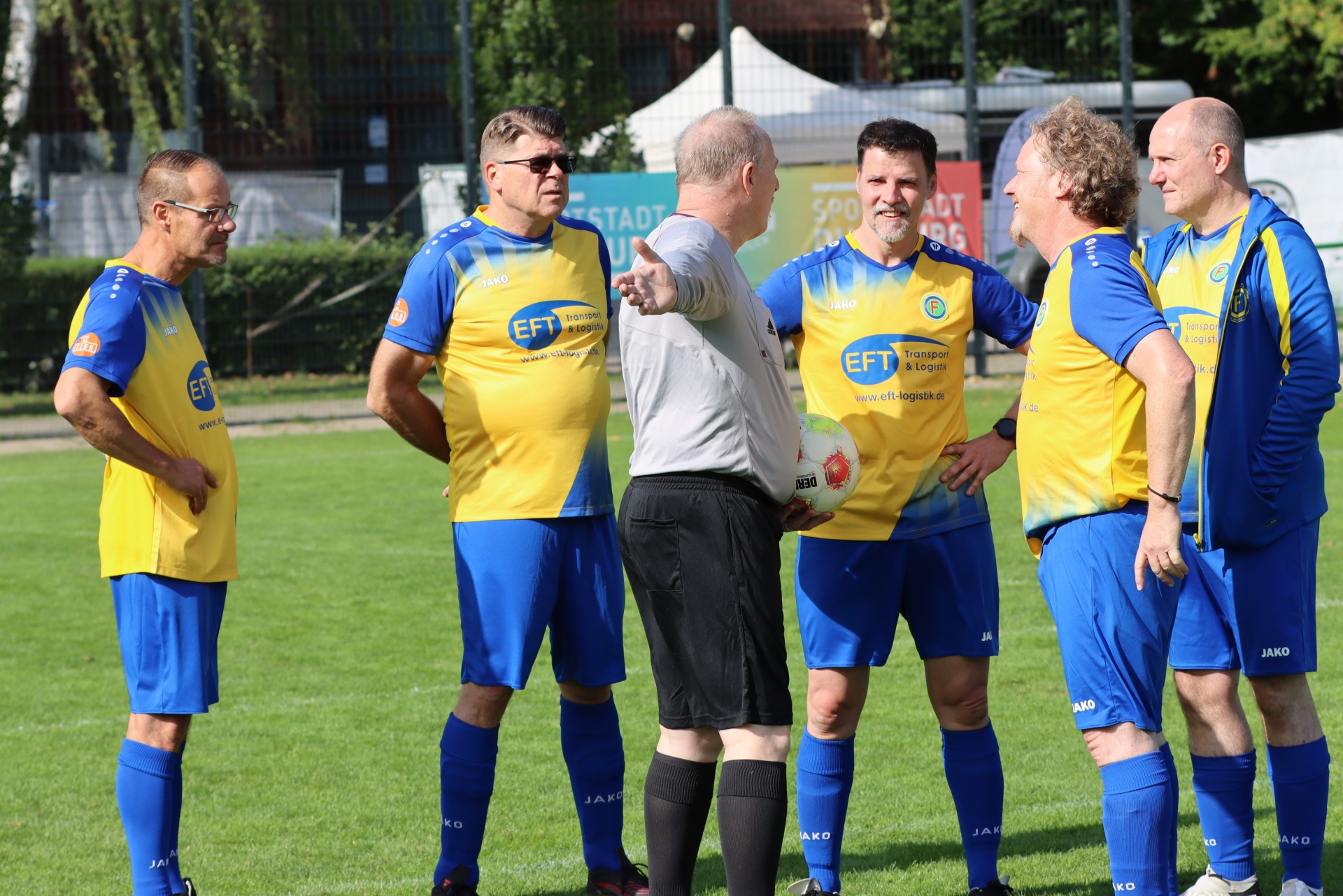[{"clang_id":1,"value":"Fußballteam in gelben Trikots und blauen Shorts steht auf grünem Rasen, diskutiert mit einem Mann im grauen Hemd."}]