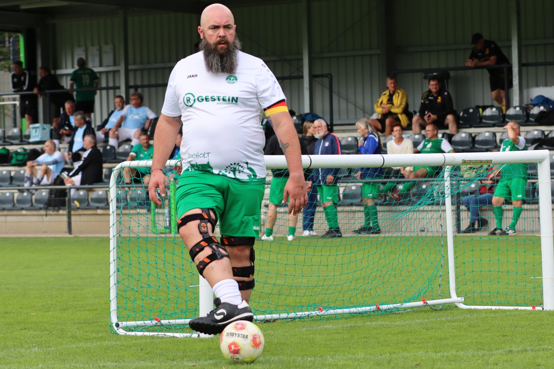 [{"clang_id":1,"value":"Fußballtorwart mit langem Bart und grünem Shorts steht vor einem weißen Fußballtor auf einem Rasenfeld. Im Hintergrund Zuschauer."}]