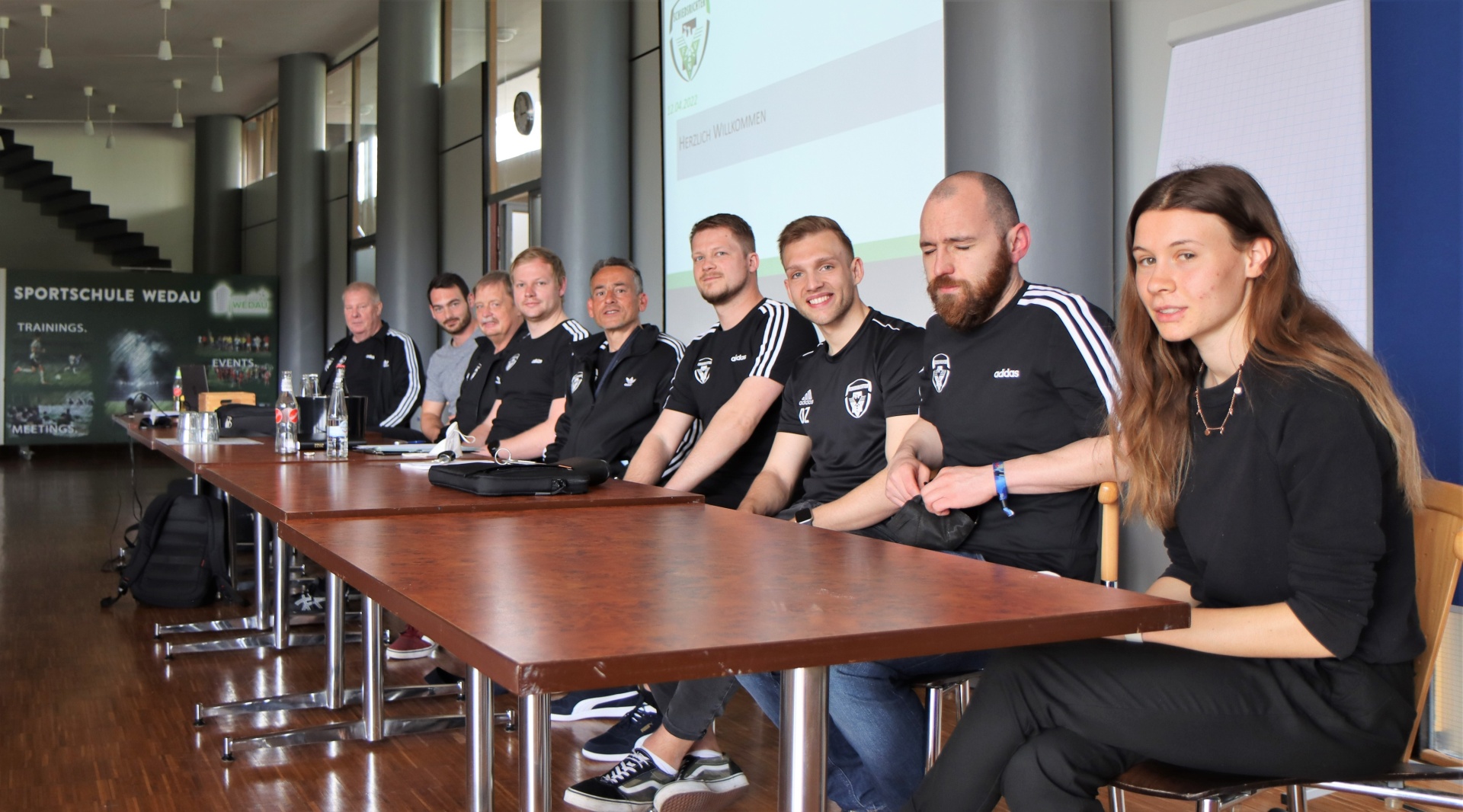 [{"clang_id":1,"value":"Gruppe von acht Personen sitzt an einem Tisch in einem hellen Raum. Sie tragen dunkle Sportkleidung. Im Hintergrund eine Präsentation und ein Banner mit der Aufschrift \"SPORTSCHULE WEDAU\"."}]