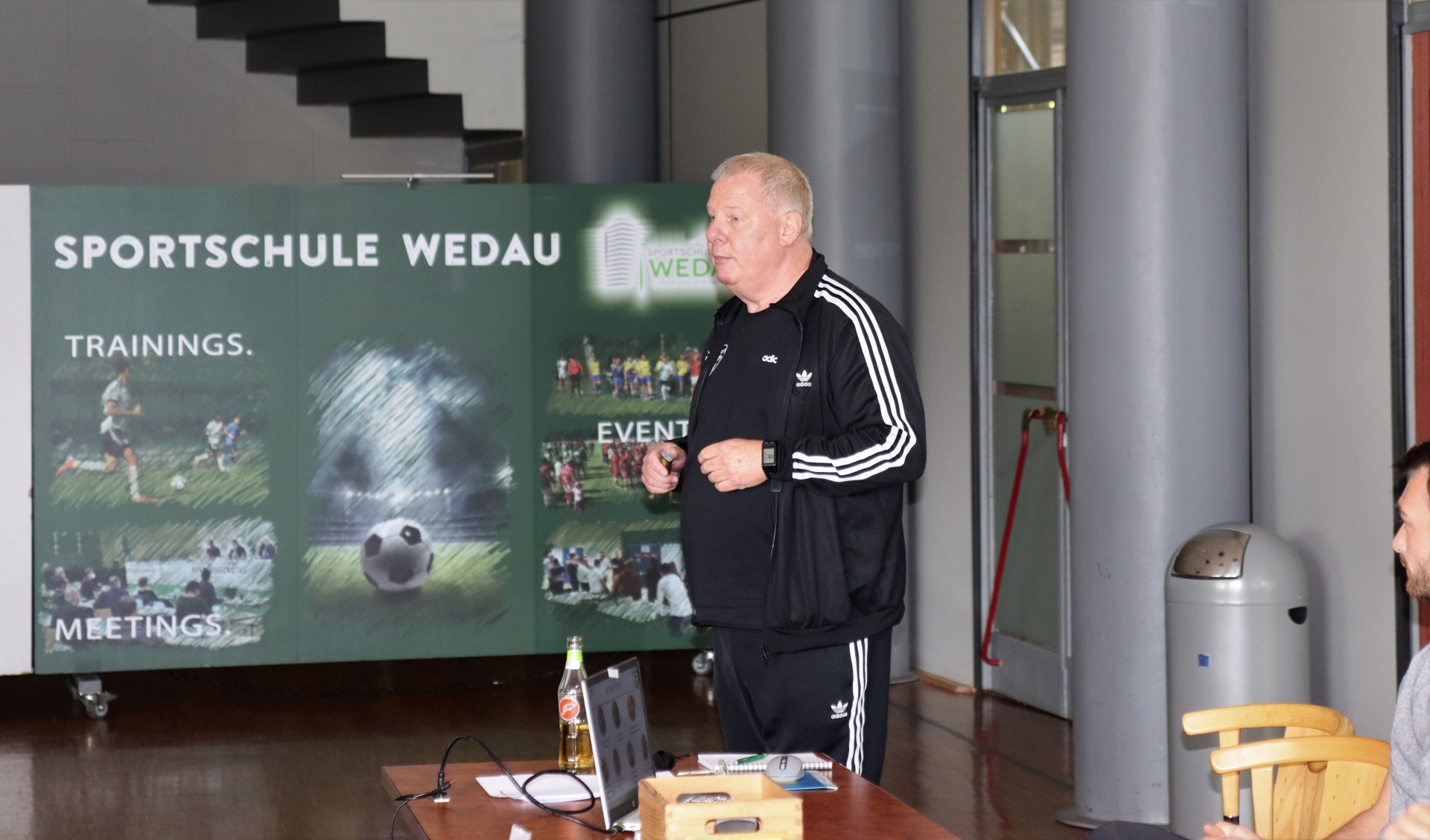 [{"clang_id":1,"value":"Mann in Sportkleidung steht vor einer Präsentation mit Sportmotiven. Im Hintergrund eine Treppe und Zuschauer auf Stühlen."}]