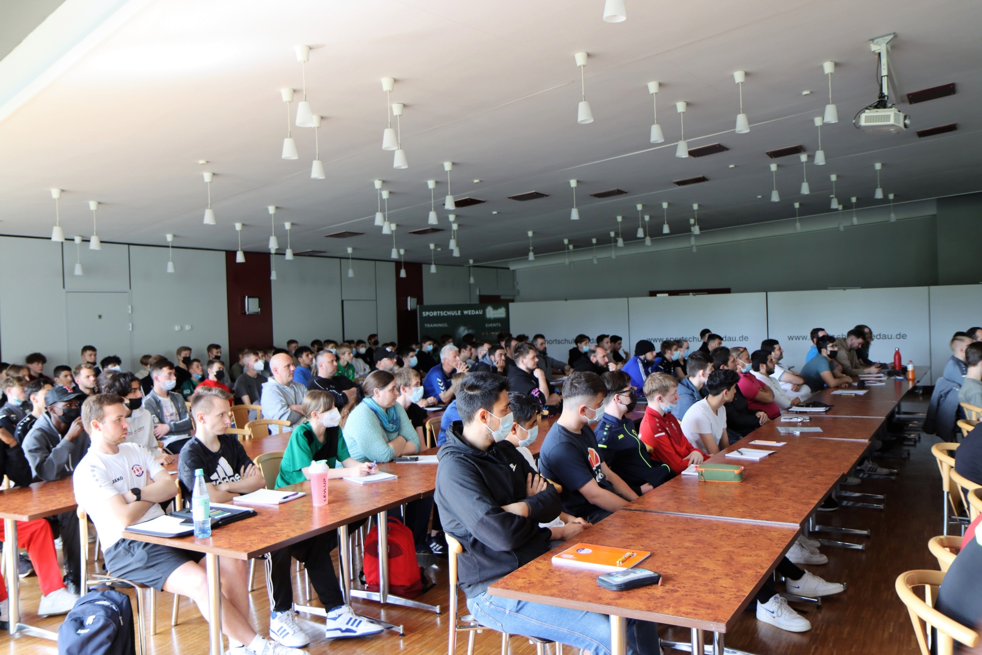 [{"clang_id":1,"value":"Großer Raum mit Sitzreihen. Viele junge Männer sitzen an Tischen, einige tragen Masken. Im Hintergrund eine Wand mit Werbeaufdruck."}]