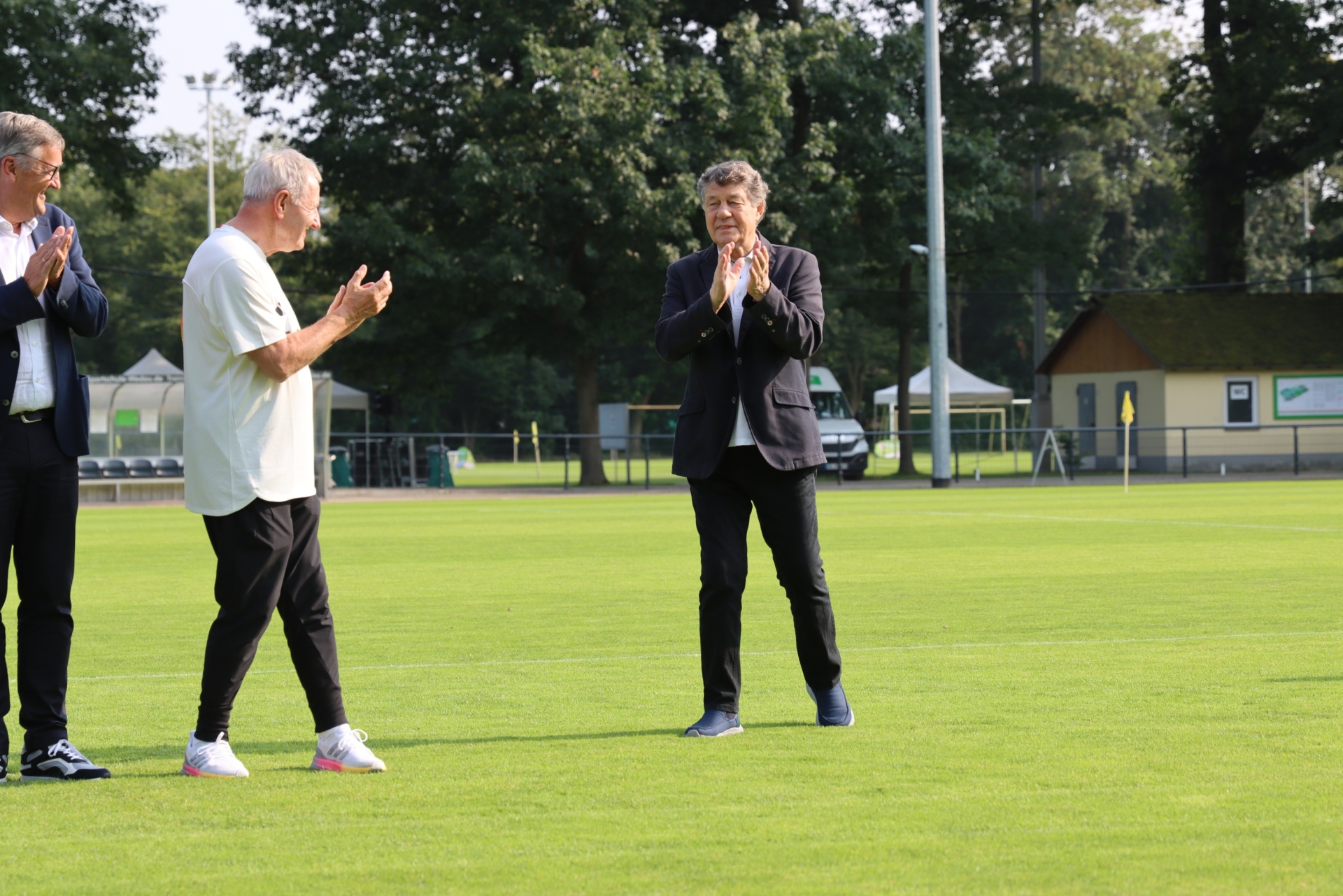 [{"clang_id":1,"value":"Drei Männer stehen auf einem Fußballfeld und klatschen in die Hände. Im Hintergrund ein Sportplatz mit Zuschauertribünen und Bäumen."}]