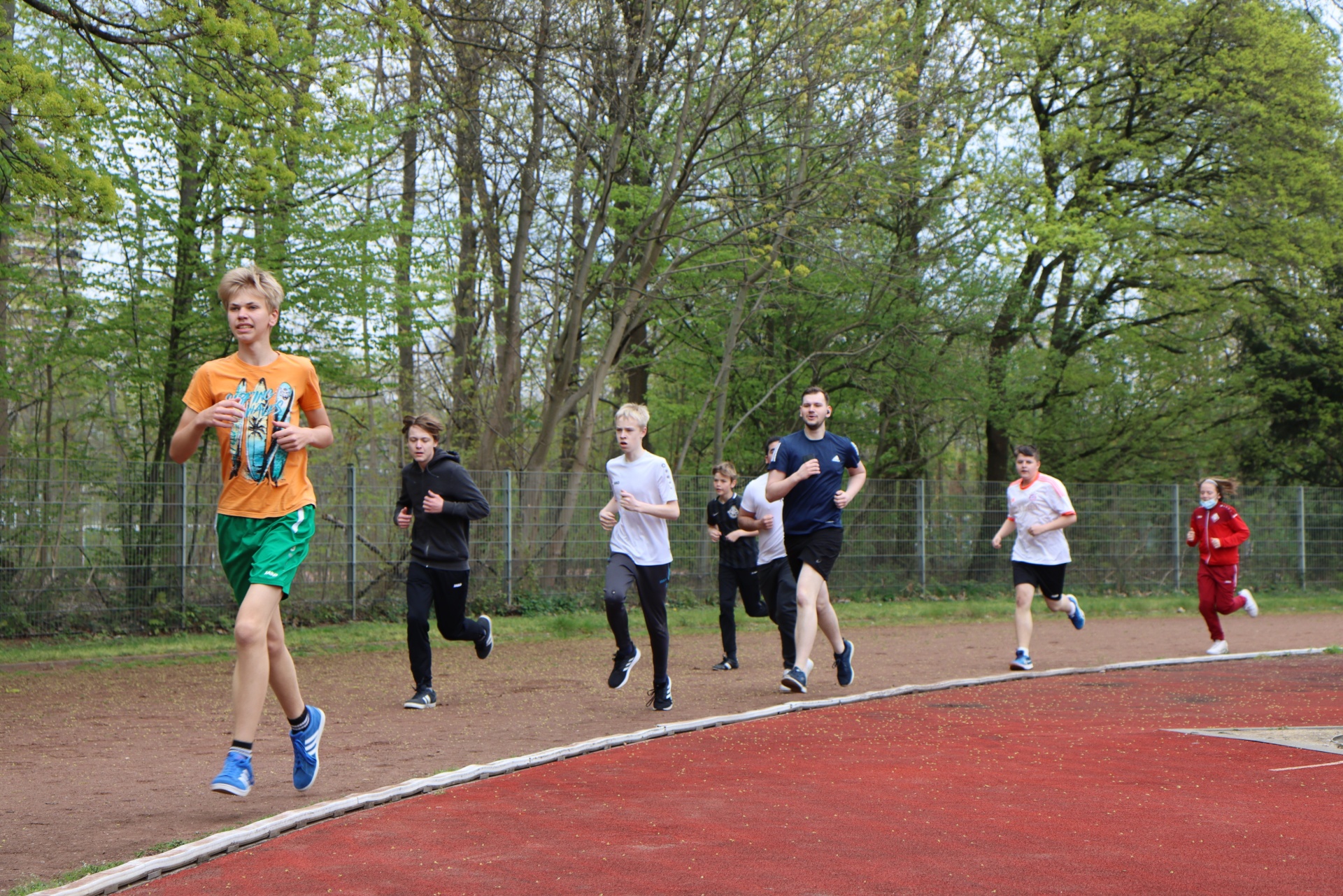 [{"clang_id":1,"value":"Gruppe von Läufern auf einer roten Tartanbahn im Freien. Vorderster Läufer trägt orangefarbenes T-Shirt und grüne Shorts. Bäume im Hintergrund."}]