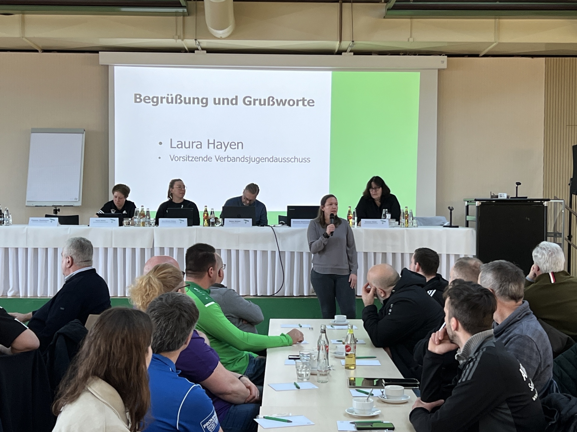 [{"clang_id":1,"value":"Versammlung in heller Halle. Rednerin am Pult, dahinter Tisch mit sieben Personen. Publikum sitzt an Tischen. Präsentation im Hintergrund mit Text \"Laura Hayen, Vorsitzende\"."}]