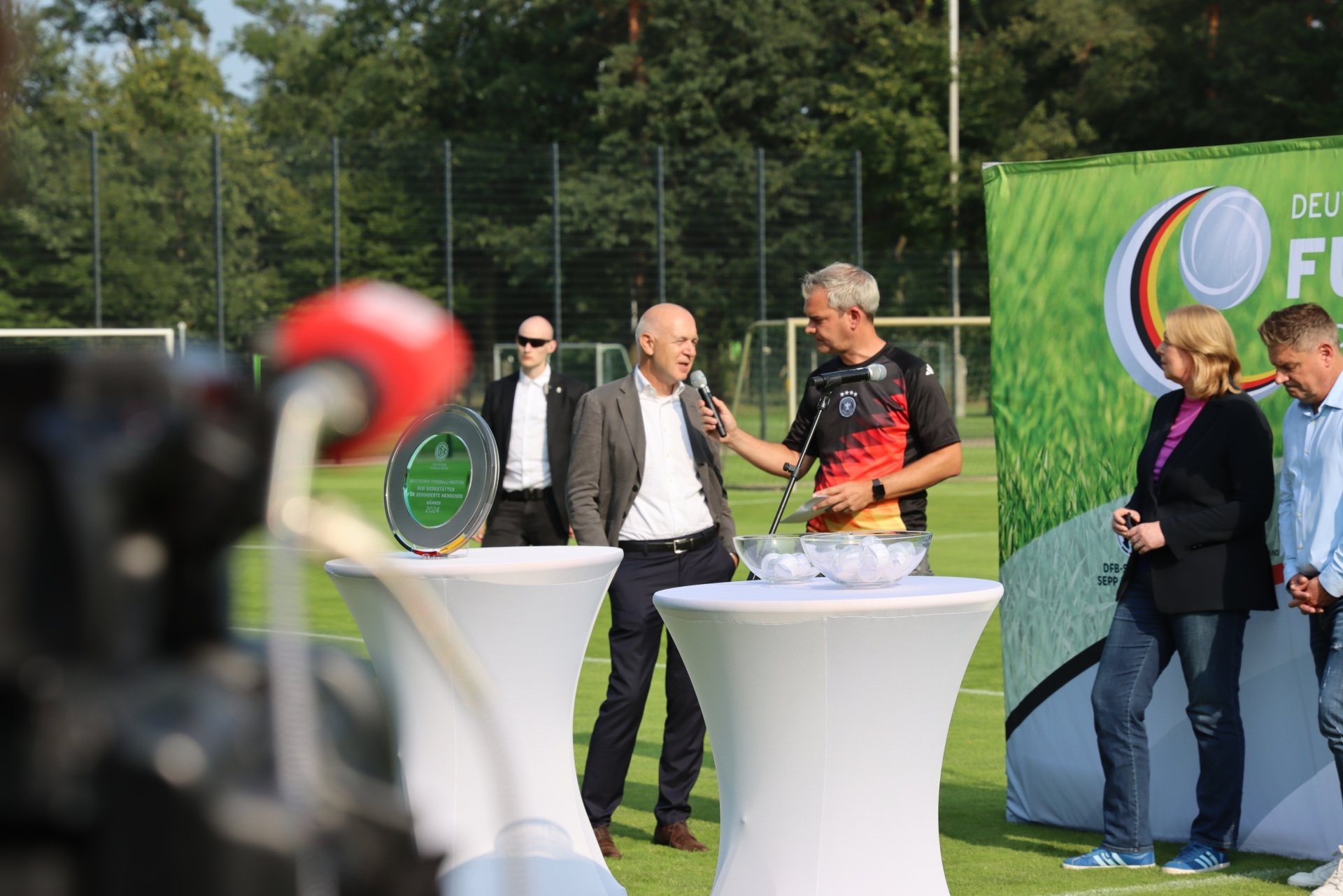 [{"clang_id":1,"value":"Fußballplatz: Männer in Anzügen halten Reden an zwei weißen Tischen. Im Hintergrund ein Banner mit deutschen Farben und Personen."}]