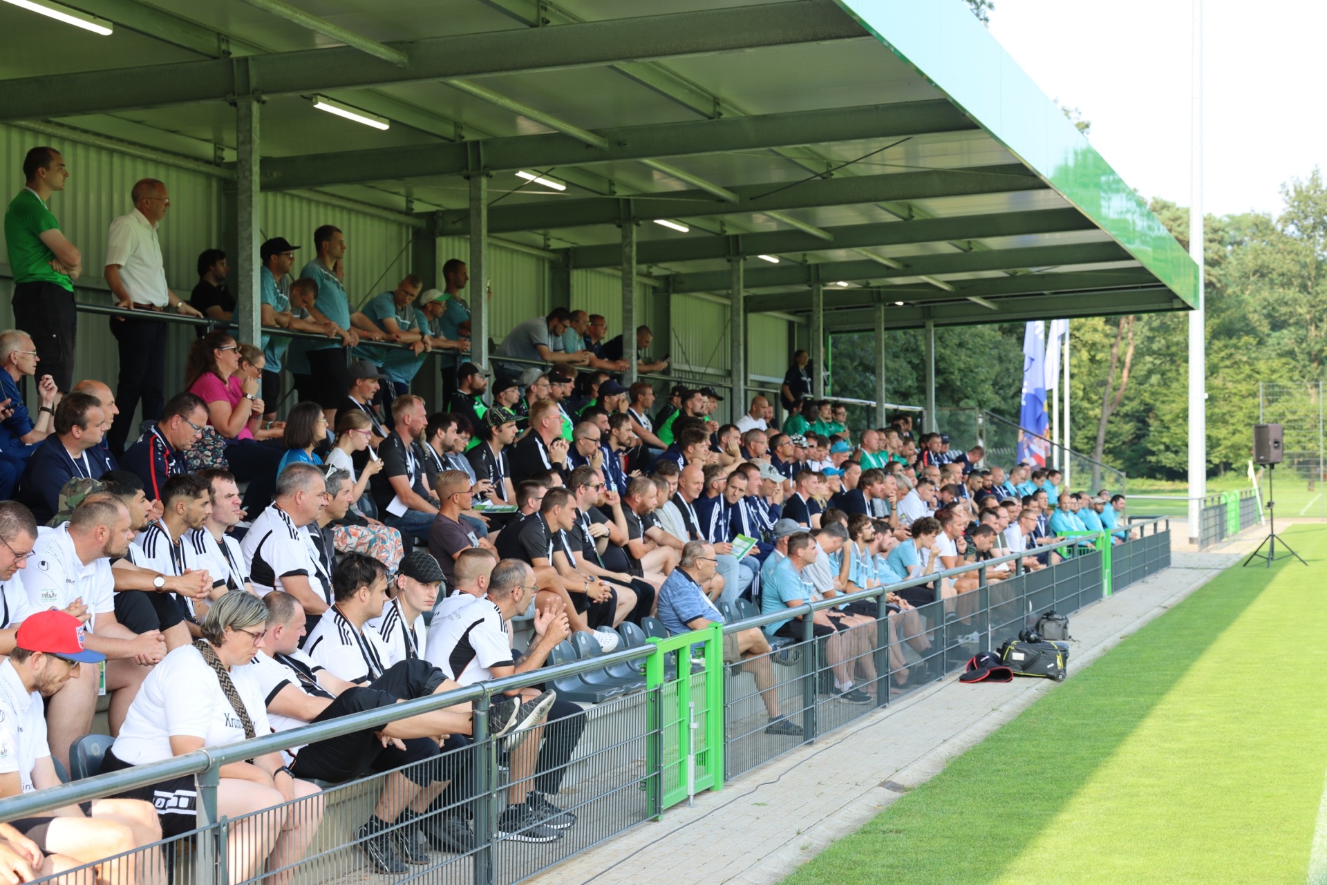 [{"clang_id":1,"value":"Zuschauerreihen an einem Sportplatz, gefüllt mit Menschen, die ein Spiel beobachten. Im Hintergrund ein Fußballfeld mit grünem Rasen."}]