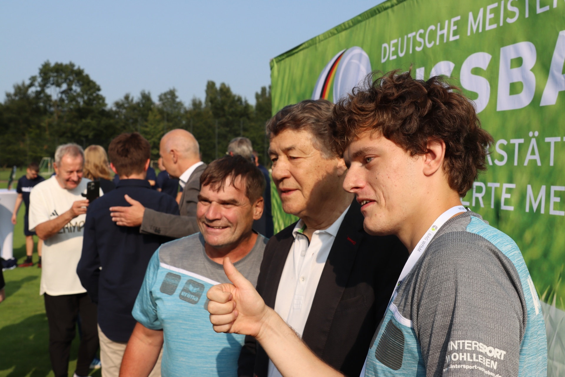 [{"clang_id":1,"value":"Gruppe von Männern im Freien vor einem Werbebanner. Ein Mann mit blauem Trikot zeigt Daumen hoch. Im Hintergrund grüne Landschaft."}]