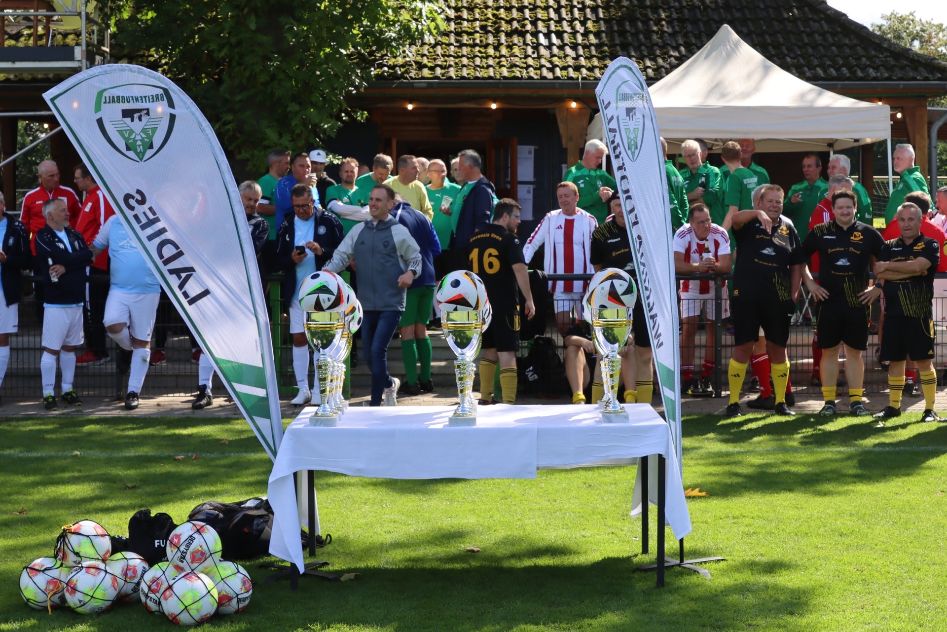 [{"clang_id":1,"value":"Fußballturnier: Gruppe Männer steht auf Rasen vor einem Tisch mit Pokalen. Im Hintergrund ein Clubhaus und Werbefahnen. Fußbälle liegen im Vordergrund."}]