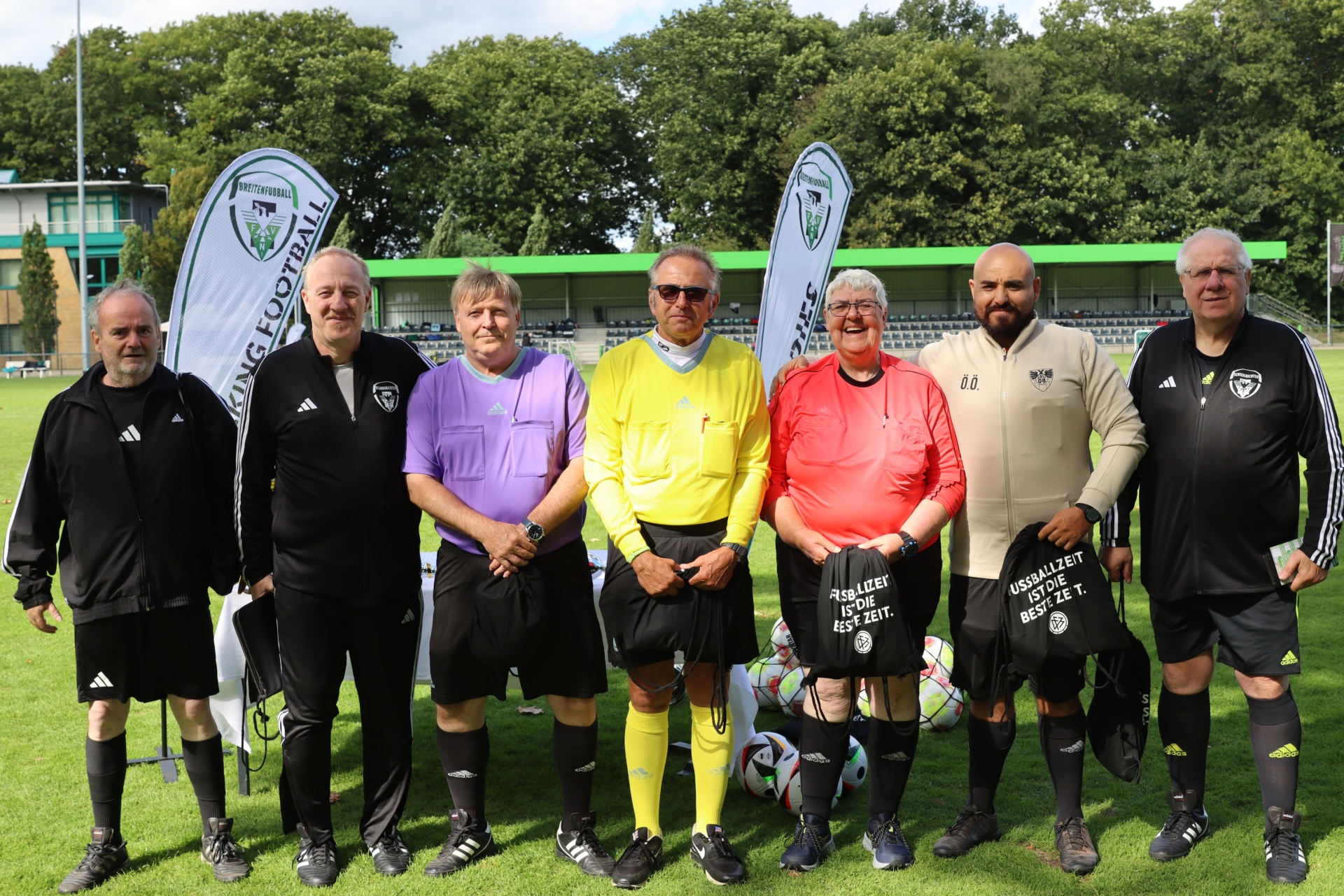 [{"clang_id":1,"value":"Gruppe von sieben Fußballschiedsrichtern in Sportkleidung steht auf einem Fußballfeld vor grünem Hintergrund und Werbebanden."}]