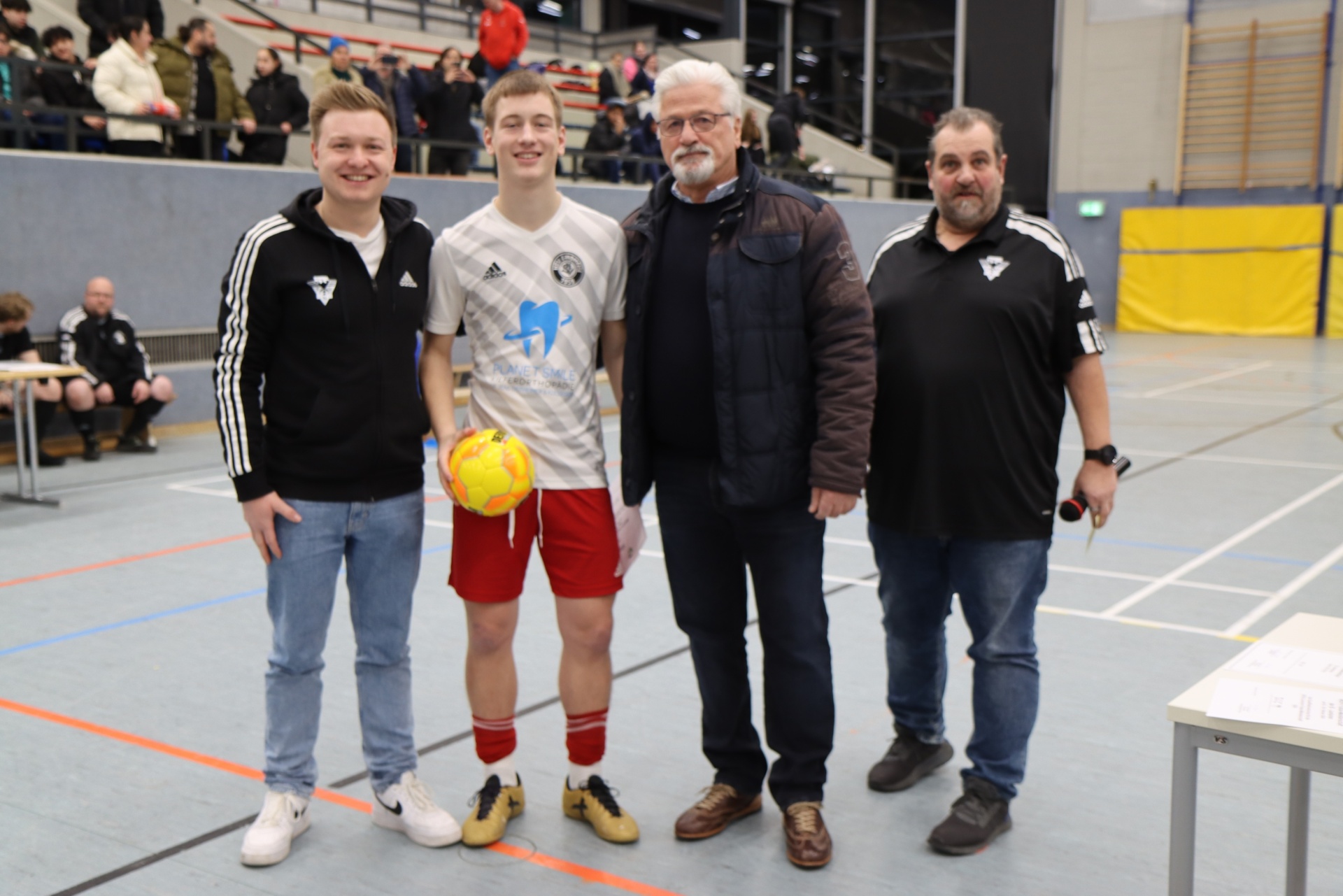 [{"clang_id":1,"value":"Vier Männer stehen in einer Sporthalle. Ein Teenager hält einen Fußball. Im Hintergrund sind Zuschauer und Sportausrüstung zu sehen."}]