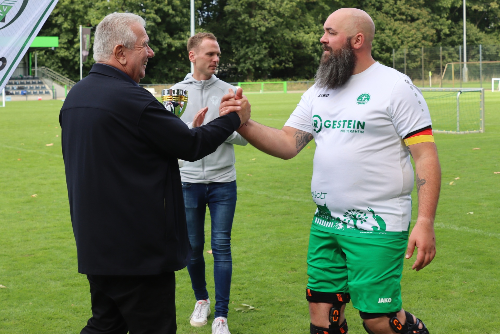 [{"clang_id":1,"value":"Zwei Männer schütteln sich die Hand auf einem Fußballfeld. Links ein älterer Mann in Anzug, rechts ein Fußballspieler mit grüner Ausrüstung. Im Hintergrund ein weiterer Mann."}]