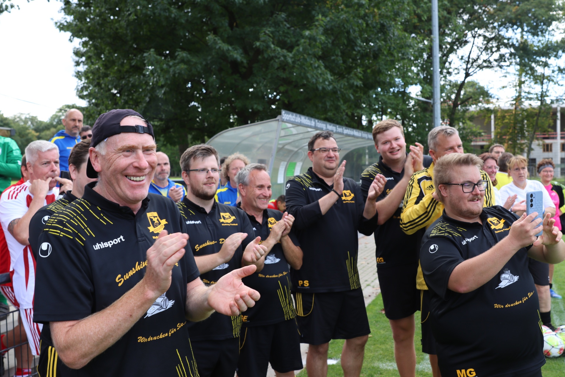 [{"clang_id":1,"value":"Fußballteam in schwarzen und gelben Trainingsanzügen applaudiert am Spielfeldrand. Im Hintergrund ein Gebäude und Zuschauer."}]