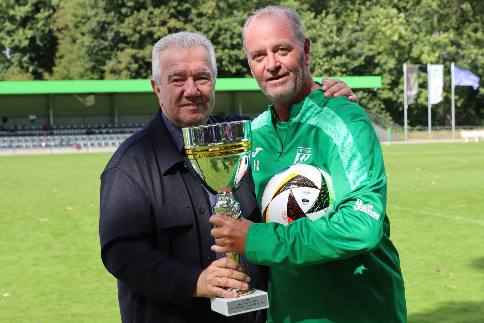 [{"clang_id":1,"value":"Zwei Männer stehen lächelnd vor einer Fußballtribüne. Links ein Mann in schwarzem Anzug, der eine goldene Trophäe hält. Rechts ein Mann in grüner Sportbekleidung mit Fußball in der Hand."}]
