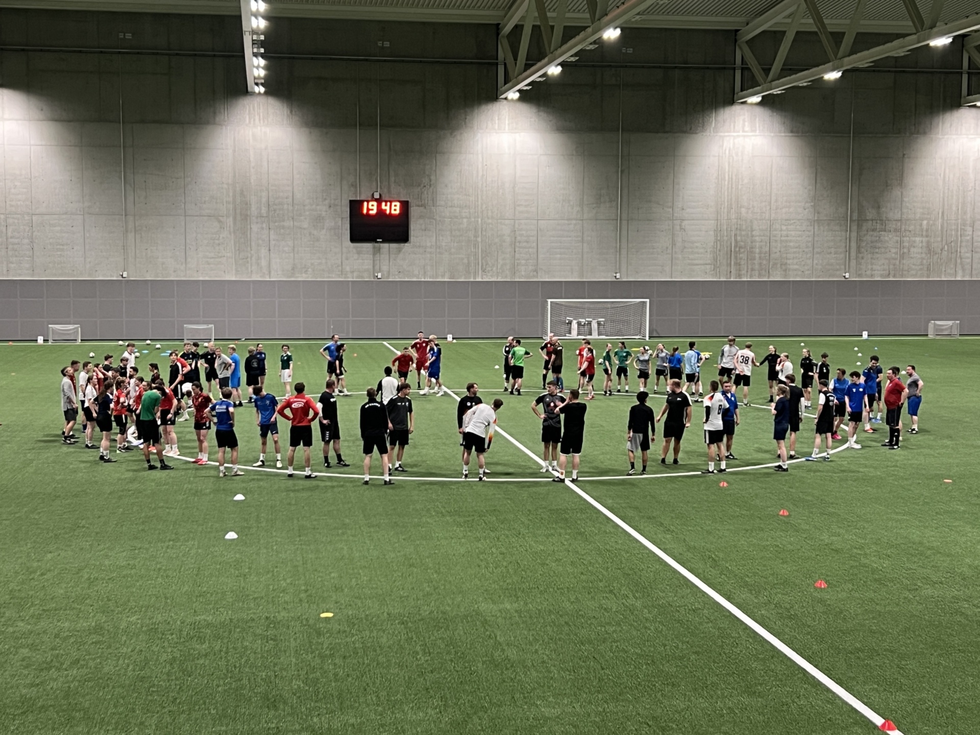 [{"clang_id":1,"value":"Fußballfeld in einer Halle. Eine große Gruppe von Spielern steht im Kreis und blickt zum Zentrum. Digitale Zeitangabe: 19:48. Ziele am Hintergrund."}]