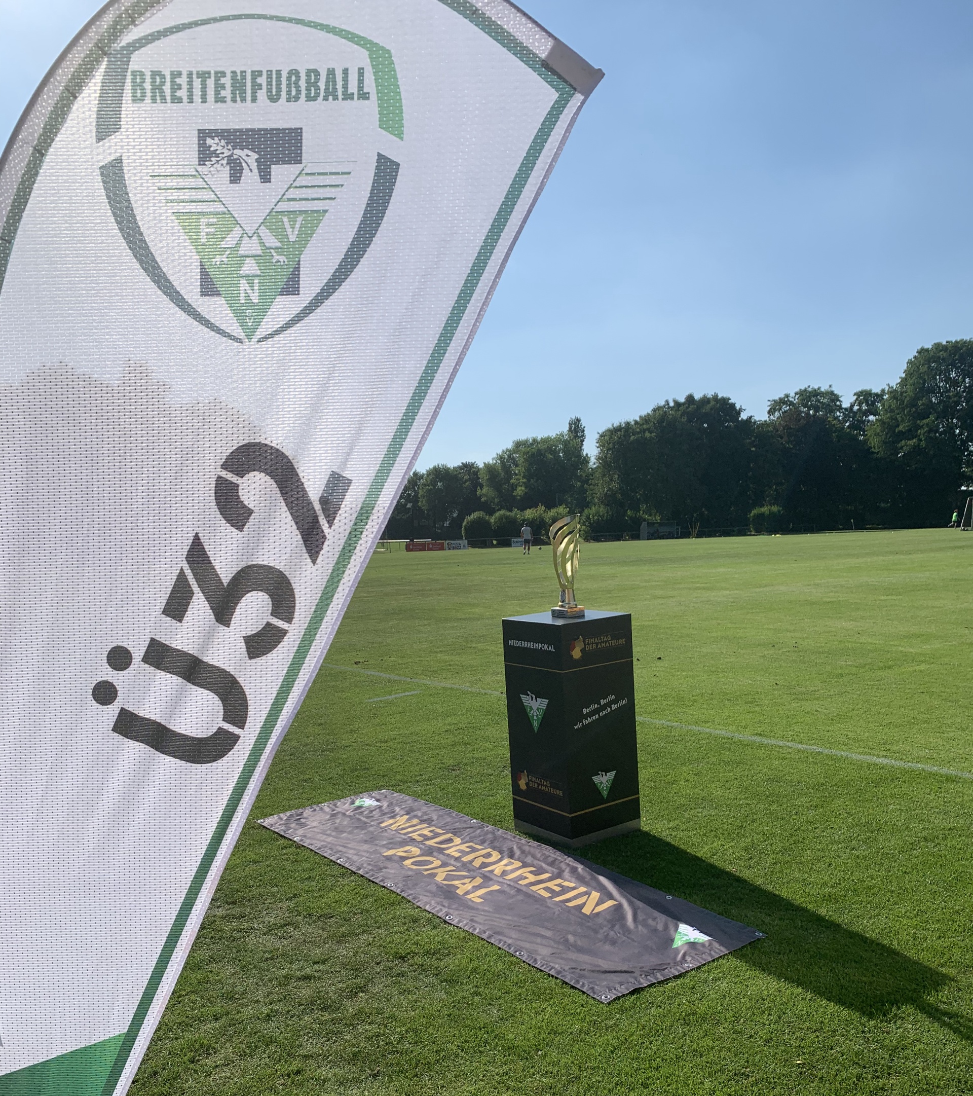 [{"clang_id":1,"value":"Fußballplatz mit Pokal im Mittelpunkt. Im Vordergrund eine Flagge mit \"Ü32\" und einem Fußballlogo. Im Hintergrund Bäume und ein blauer Himmel."}]