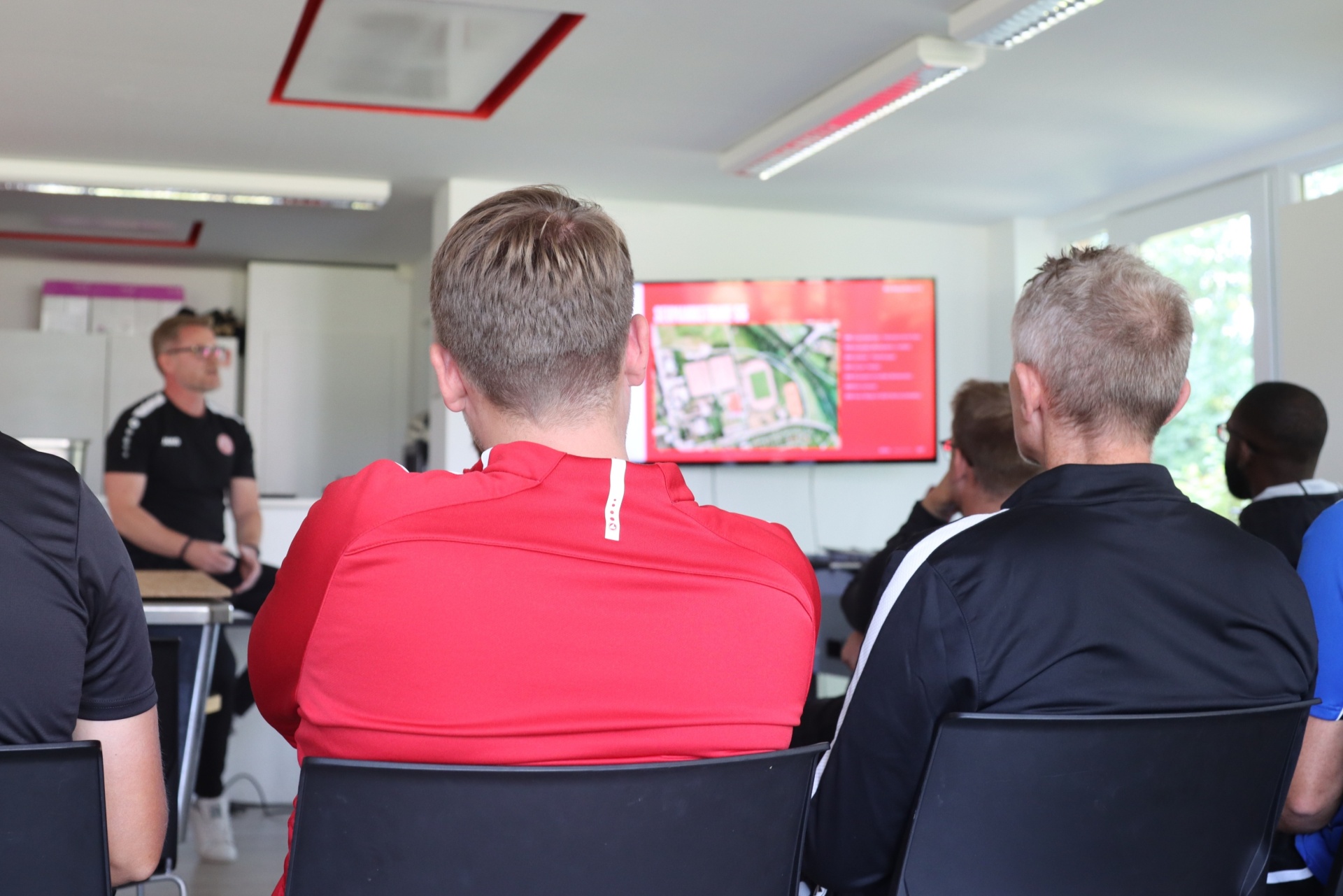 [{"clang_id":1,"value":"Klassenzimmer mit Sitzgruppe vor einer großen Leinwand. Ein Mann steht vorne und spricht, mehrere Personen sitzen ihm zugewandt."}]