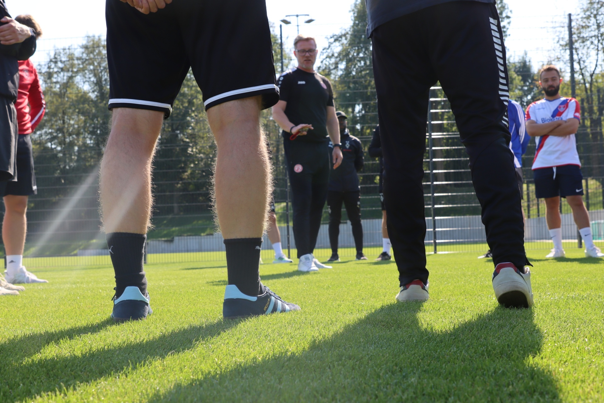 [{"clang_id":1,"value":"Fußballtraining: Zwei Spieler mit Rücken zum Betrachter stehen auf grünem Rasen. Im Hintergrund weitere Spieler und ein Trainer."}]