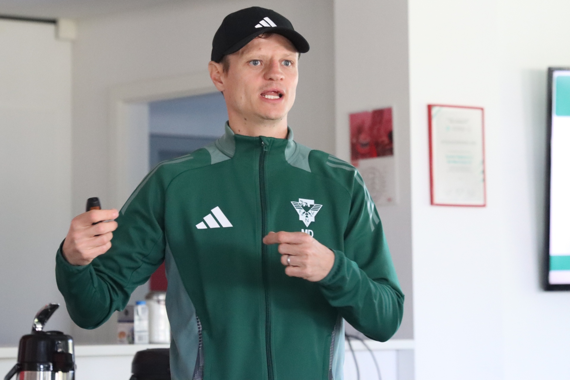 [{"clang_id":1,"value":"Mann mit heller Haut, grüner Trainingsjacke und schwarzer Kappe spricht in ein Mikrofon. Im Hintergrund eine Wand mit Bildern."}]