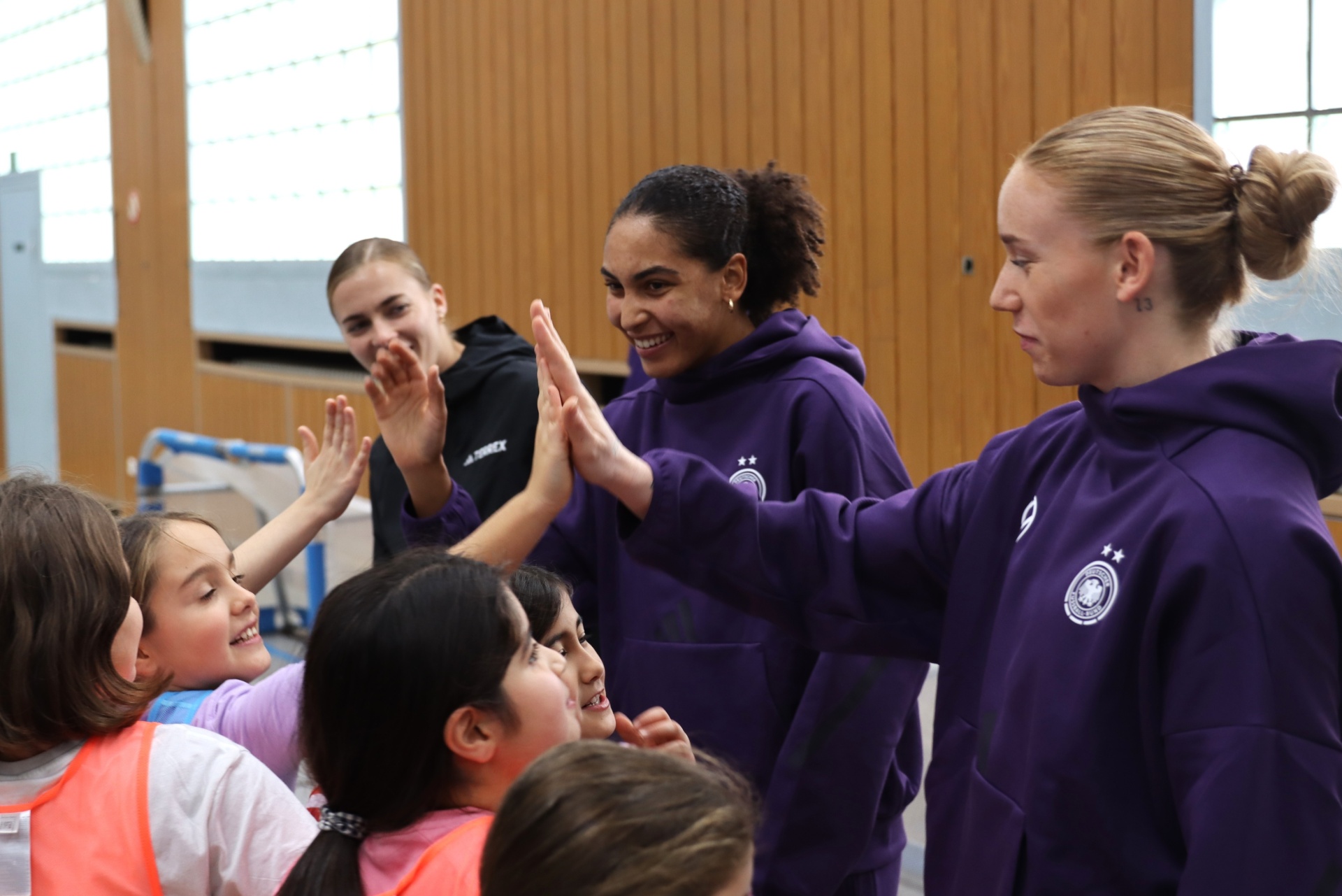 [{"clang_id":1,"value":"Drei Sportlerinnen in violetten Trainingsanzügen geben High-Fives an eine Gruppe lächelnder Kinder in einem hellen Hallensportraum."}]