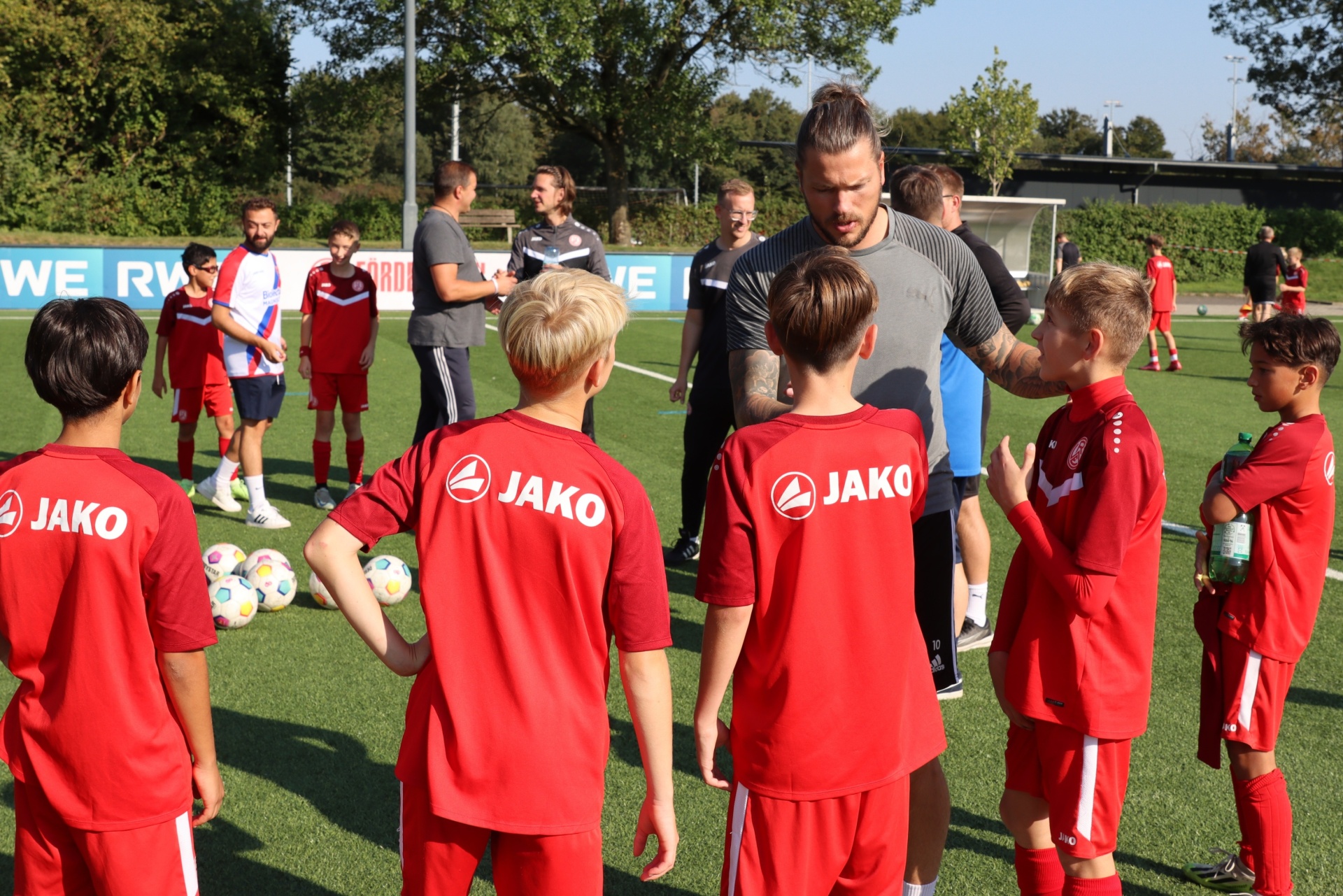 [{"clang_id":1,"value":"Fußballtraining: Eine Gruppe Kinder in roten Trikots steht um einen Erwachsenen auf einem grünen Fußballplatz. Im Hintergrund weitere Personen und ein Zaun."}]
