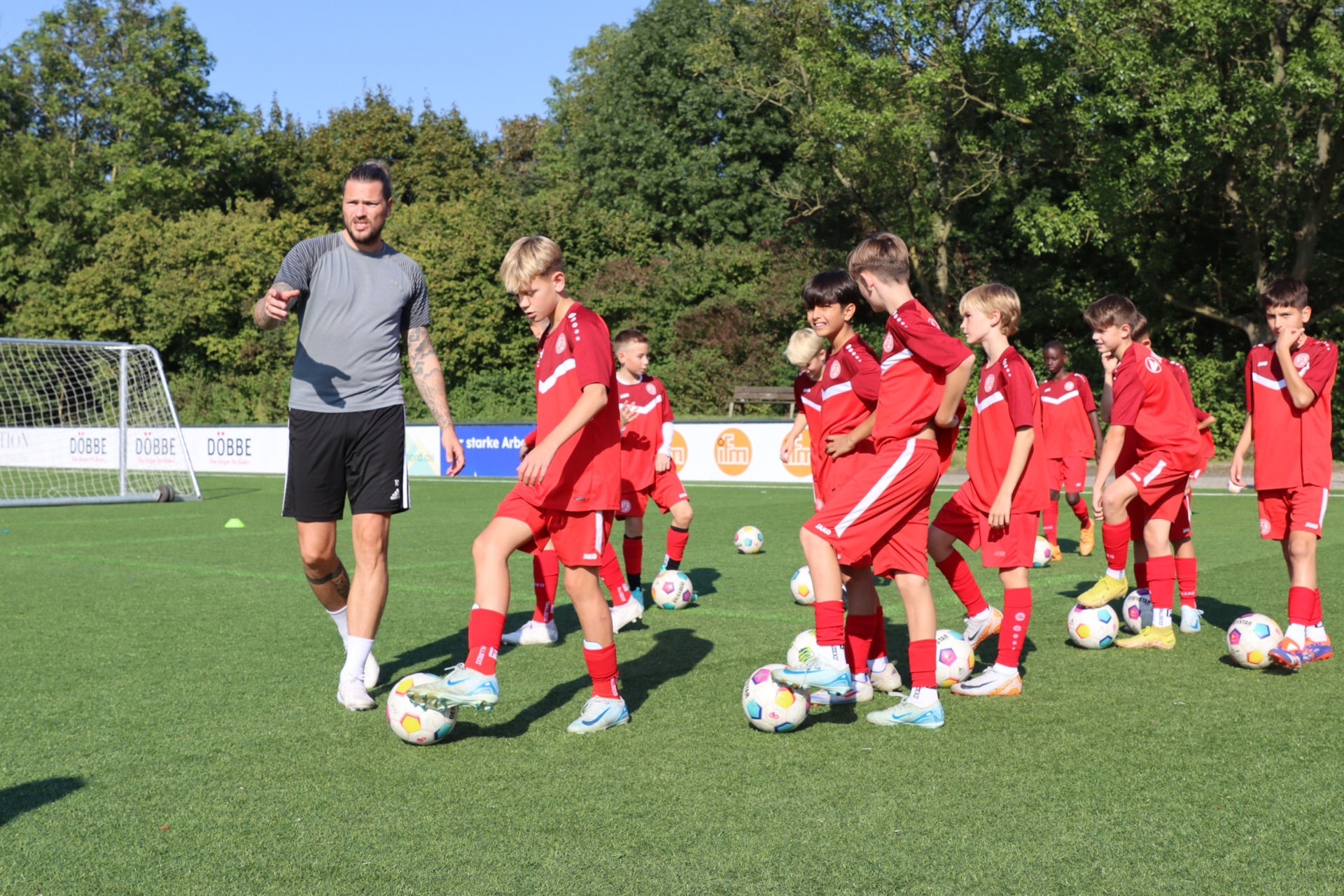 [{"clang_id":1,"value":"Fußballtrainer und Jugendmannschaft im Training auf grünem Rasenplatz. Die Kinder in roten Trikots üben mit Fußbällen. Im Hintergrund Bäume."}]