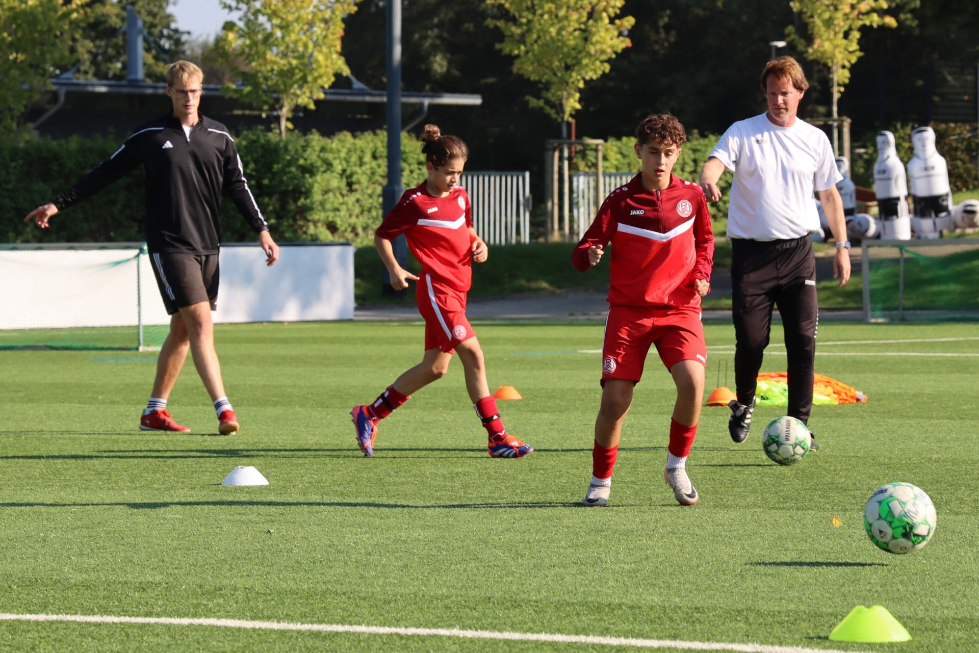 [{"clang_id":1,"value":"Fußballtraining: Zwei Jugendliche in roten Trikots dribbeln auf grünem Rasen, angeleitet von zwei erwachsenen Trainern. Trainingsmarkierungen gelb und weiß."}]