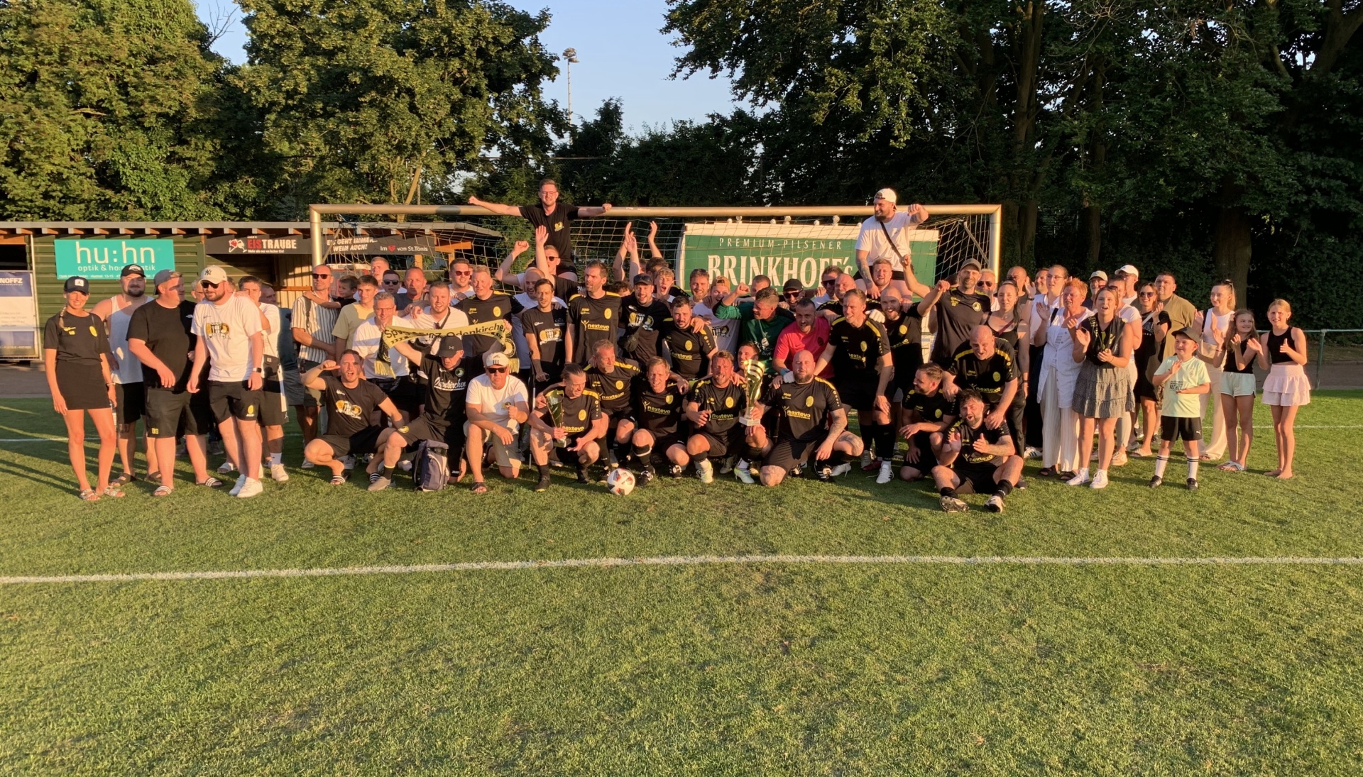 [{"clang_id":1,"value":"Fußballmannschaft in schwarzen und gelben Trainingsanzügen posiert auf grünem Rasen vor einem Fußballtor und Zuschauern unter Bäumen."}]