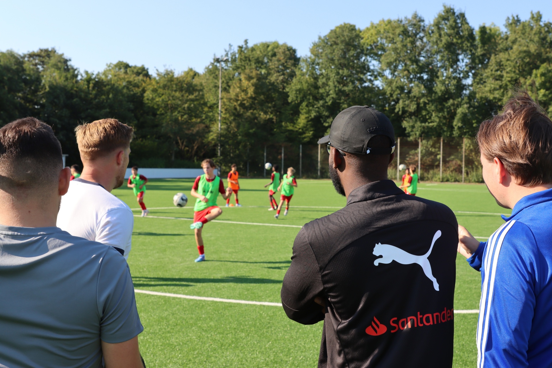 [{"clang_id":1,"value":"Fußballfeld mit spielenden Kindern. Im Vordergrund drei Männer, die dem Spiel zuschauen. Einer trägt eine schwarze Jacke mit PUMA- und Santander-Logo."}]