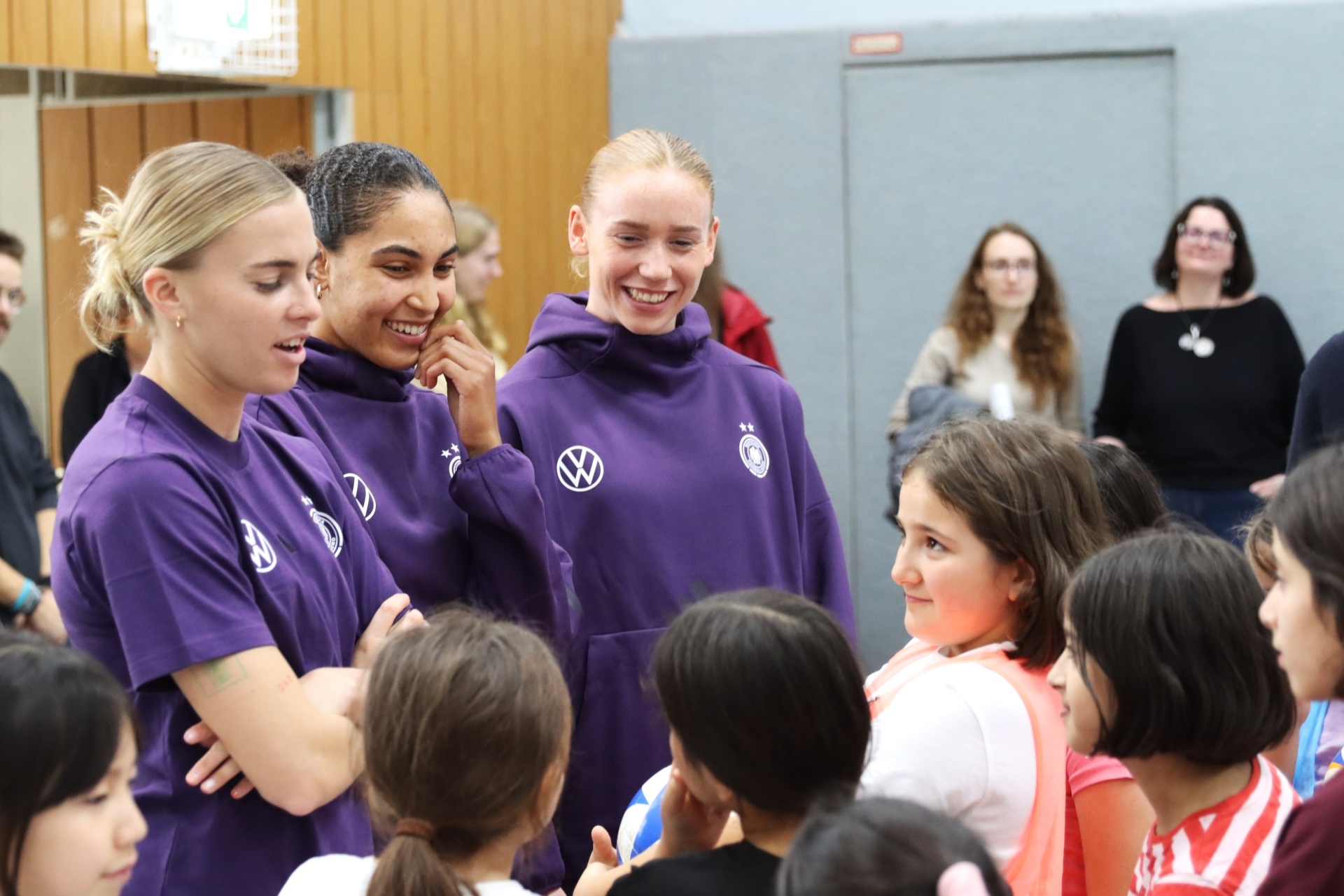 [{"clang_id":1,"value":"Drei Fußballspielerinnen in violetten Trainingsanzügen interagieren lächelnd mit einer Gruppe junger Mädchen. Im Hintergrund weitere Personen in einem Innenraum."}]