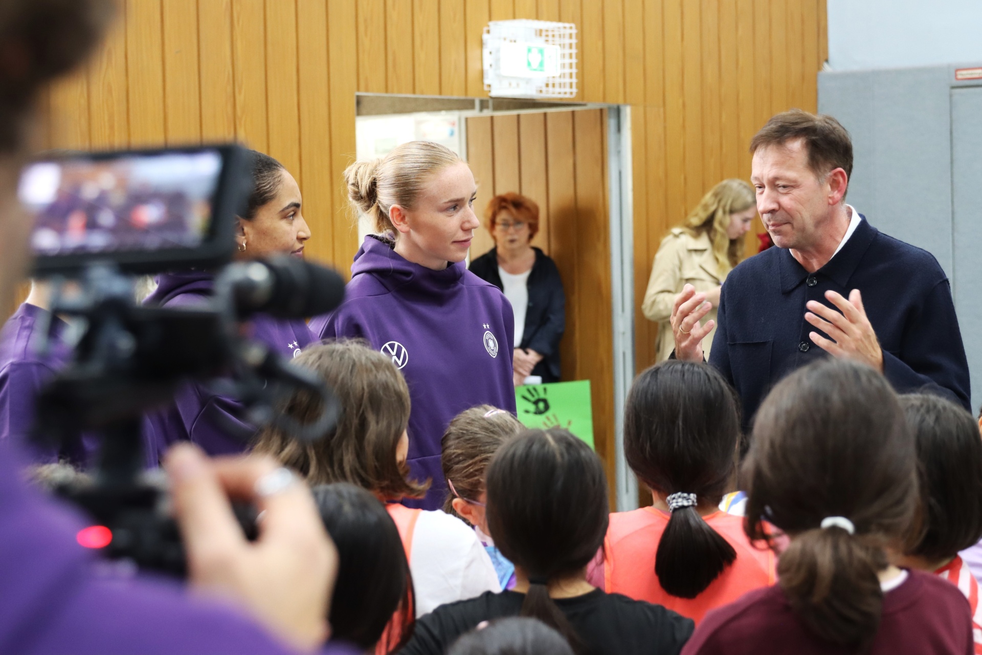 [{"clang_id":1,"value":"Gruppe von Menschen im Innenraum, darunter zwei Sportlerinnen in violetten Trainingsanzügen und ein Mann im Jackett, der spricht. Im Vordergrund eine Kamera und Kinder."}]