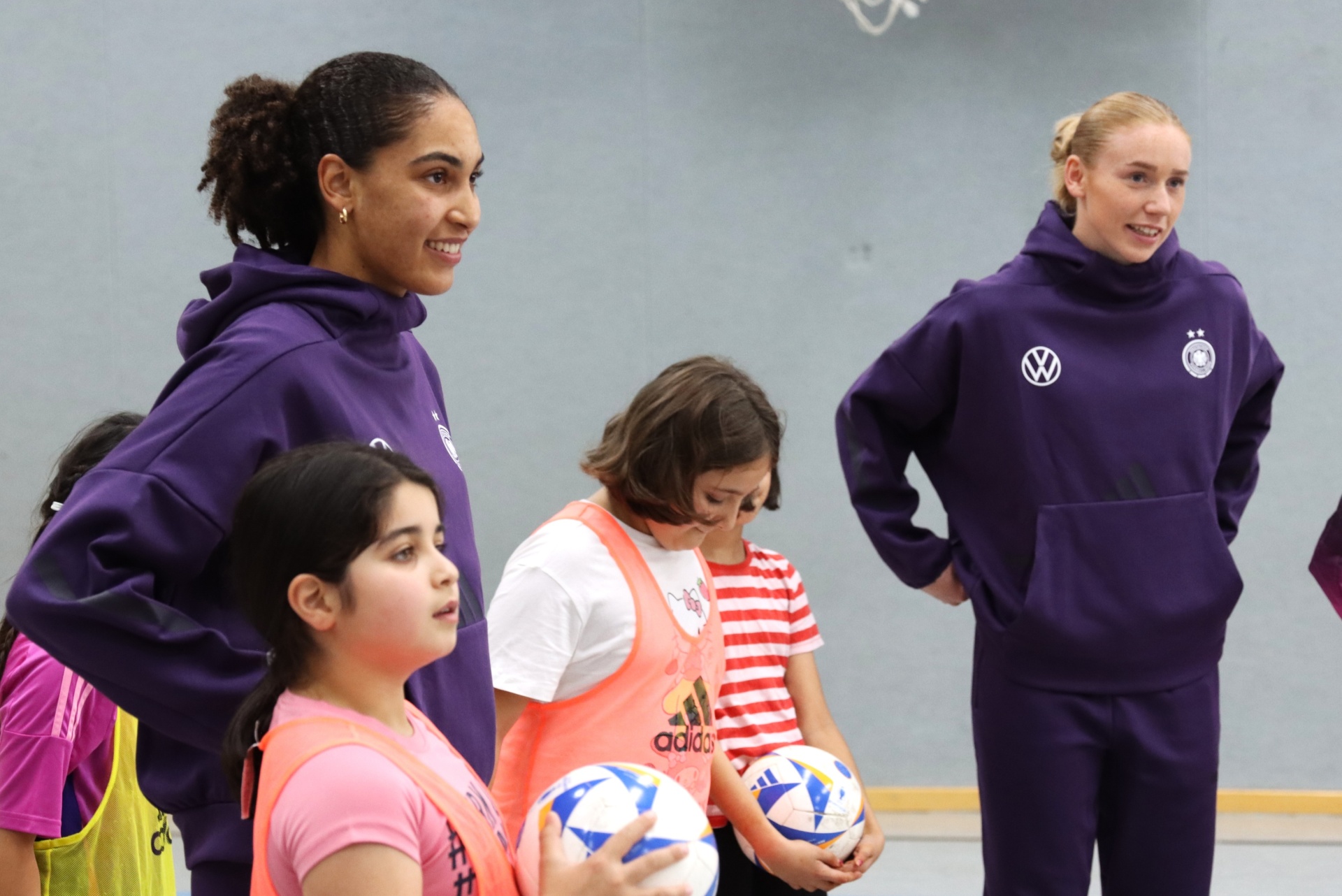 [{"clang_id":1,"value":"Zwei erwachsene Frauen und drei Kinder stehen in einer Sporthalle. Eine Frau trägt ein violettes Trainingsoutfit, die andere ein violettes Sweatshirt mit Logo. Die Kinder halten Fußbälle."}]