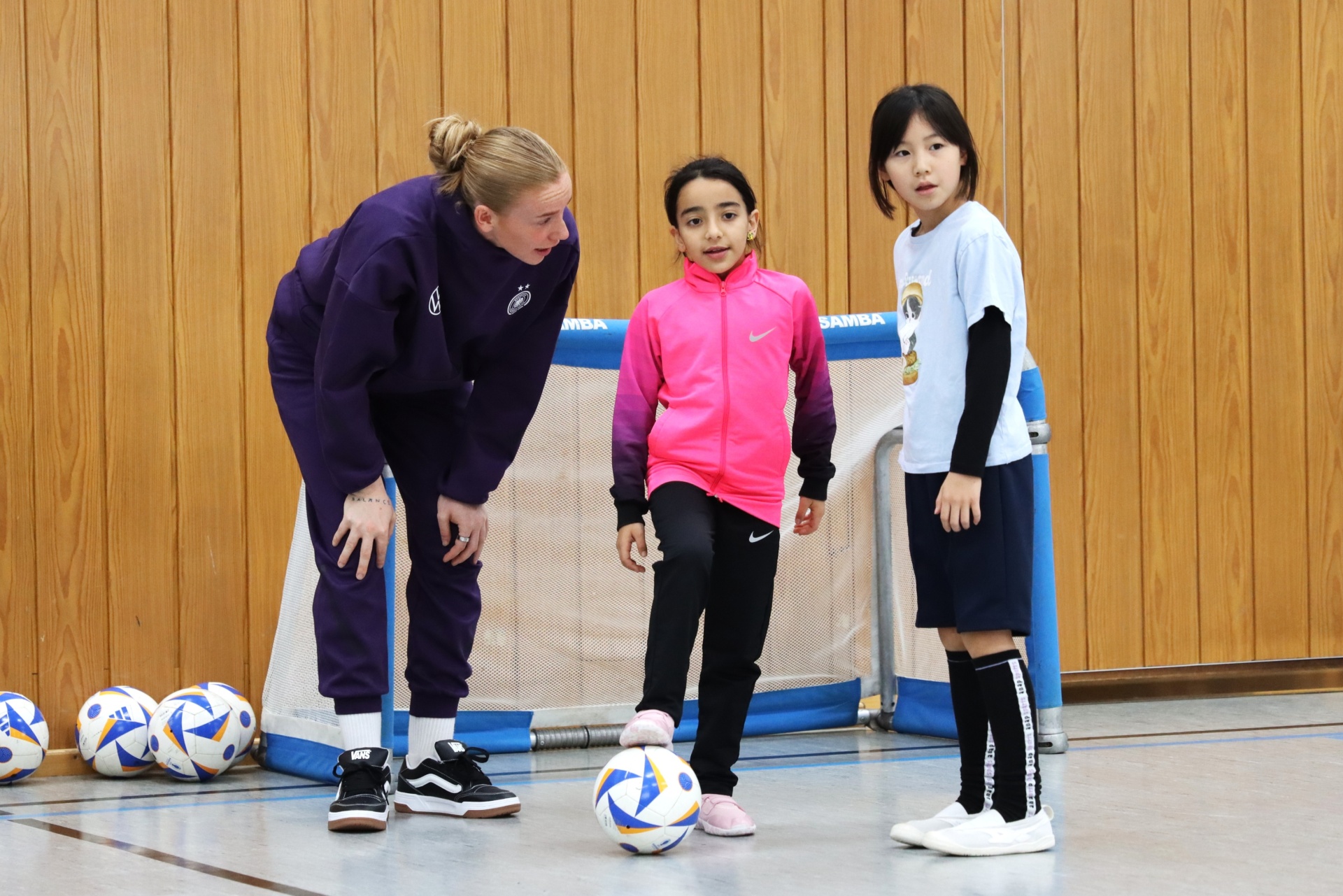 [{"clang_id":1,"value":"Drei Personen in Sporthalle: Erwachsene trainiert zwei Kinder Fußball. Viele Fußbälle liegen am Boden. Holzvertäfelung im Hintergrund."}]