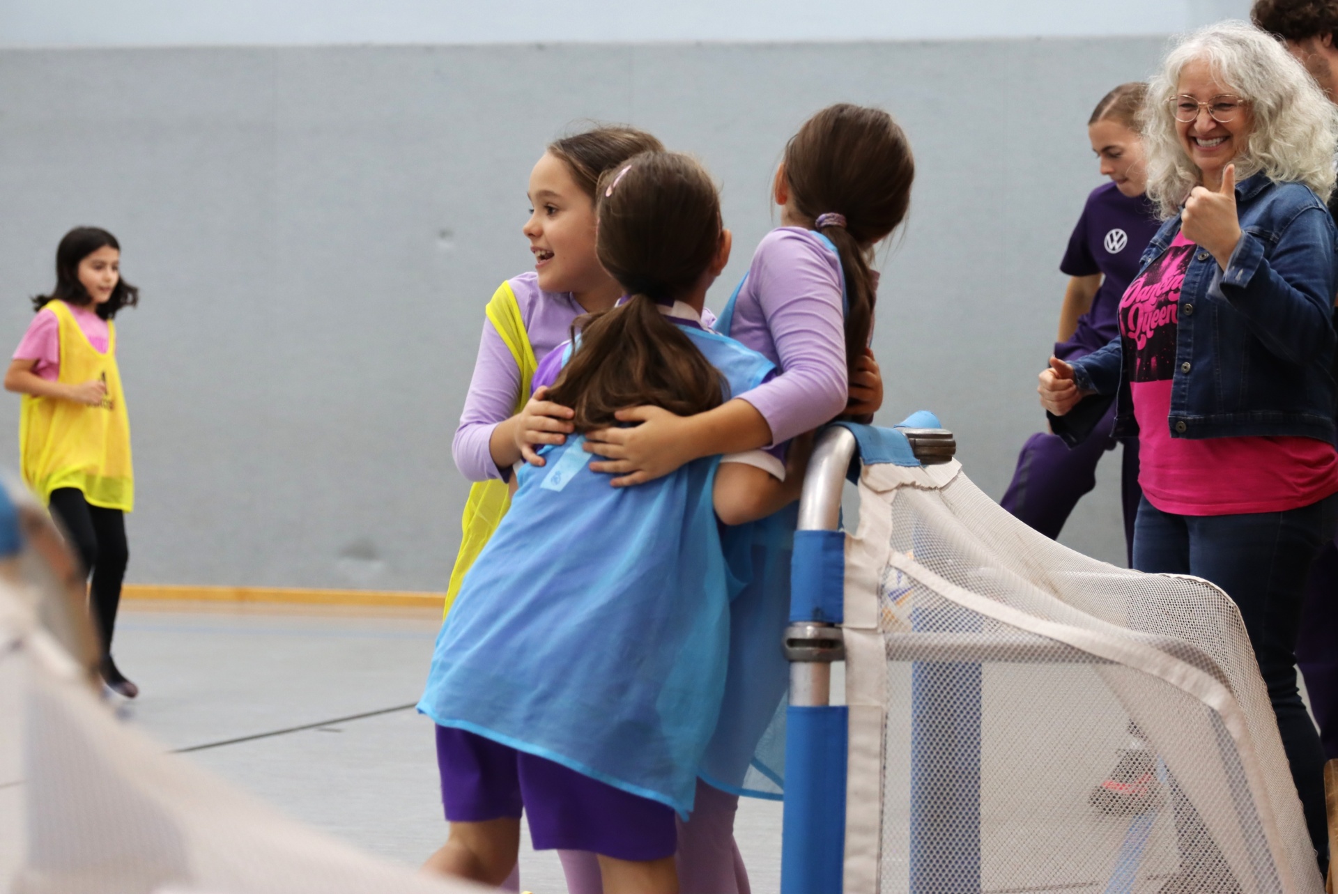 [{"clang_id":1,"value":"Schülerinnen umarmen sich freudig nach einem Sportspiel vor einem grauen Hintergrund. Im Hintergrund stehen weitere Kinder und eine erwachsene Person. Ein Hockeytor ist teilweise zu sehen."}]