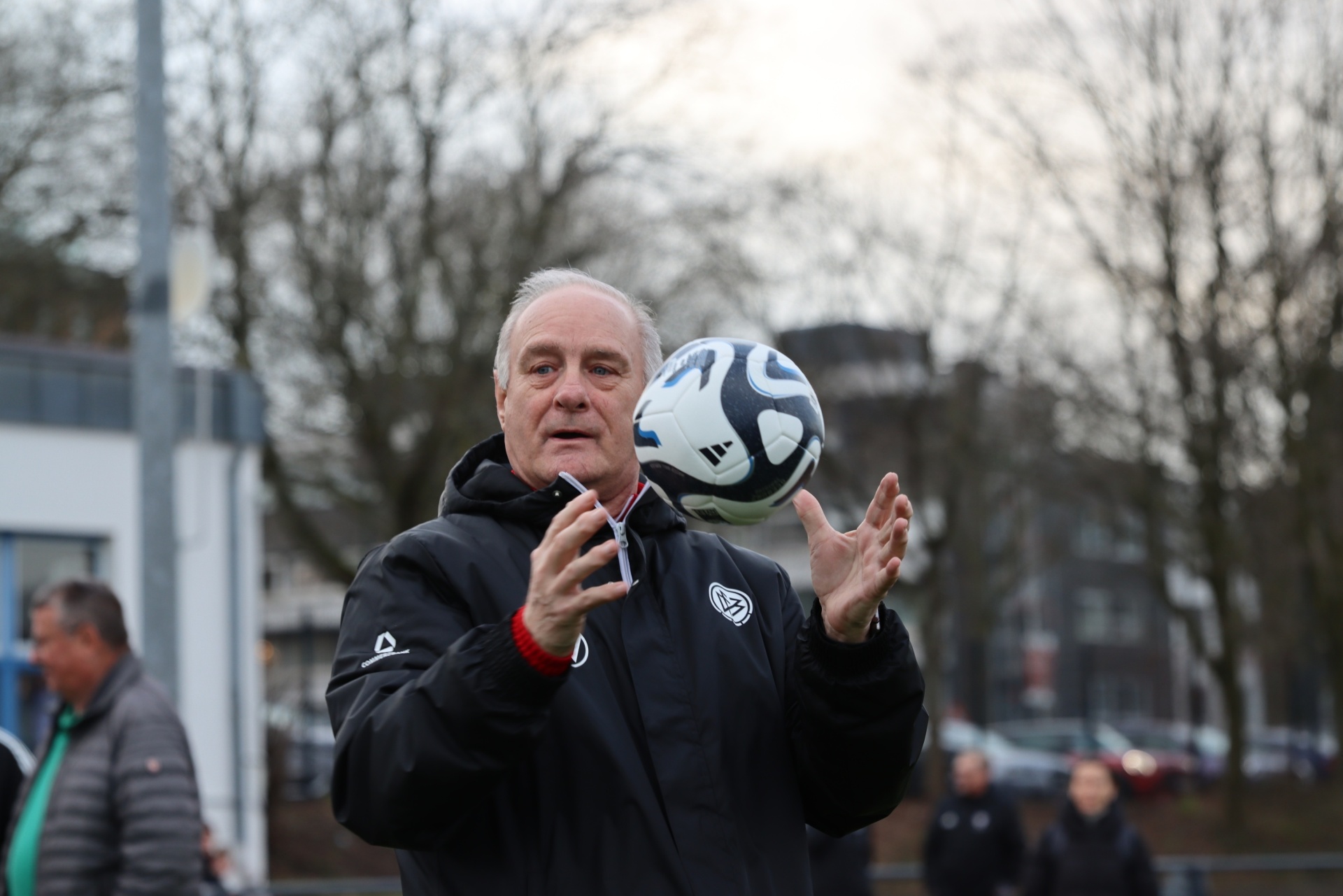 [{"clang_id":1,"value":"Mann jongliert mit einem Fußball. Er trägt eine dunkle Jacke und steht auf einem Sportplatz mit Bäumen im Hintergrund."}]