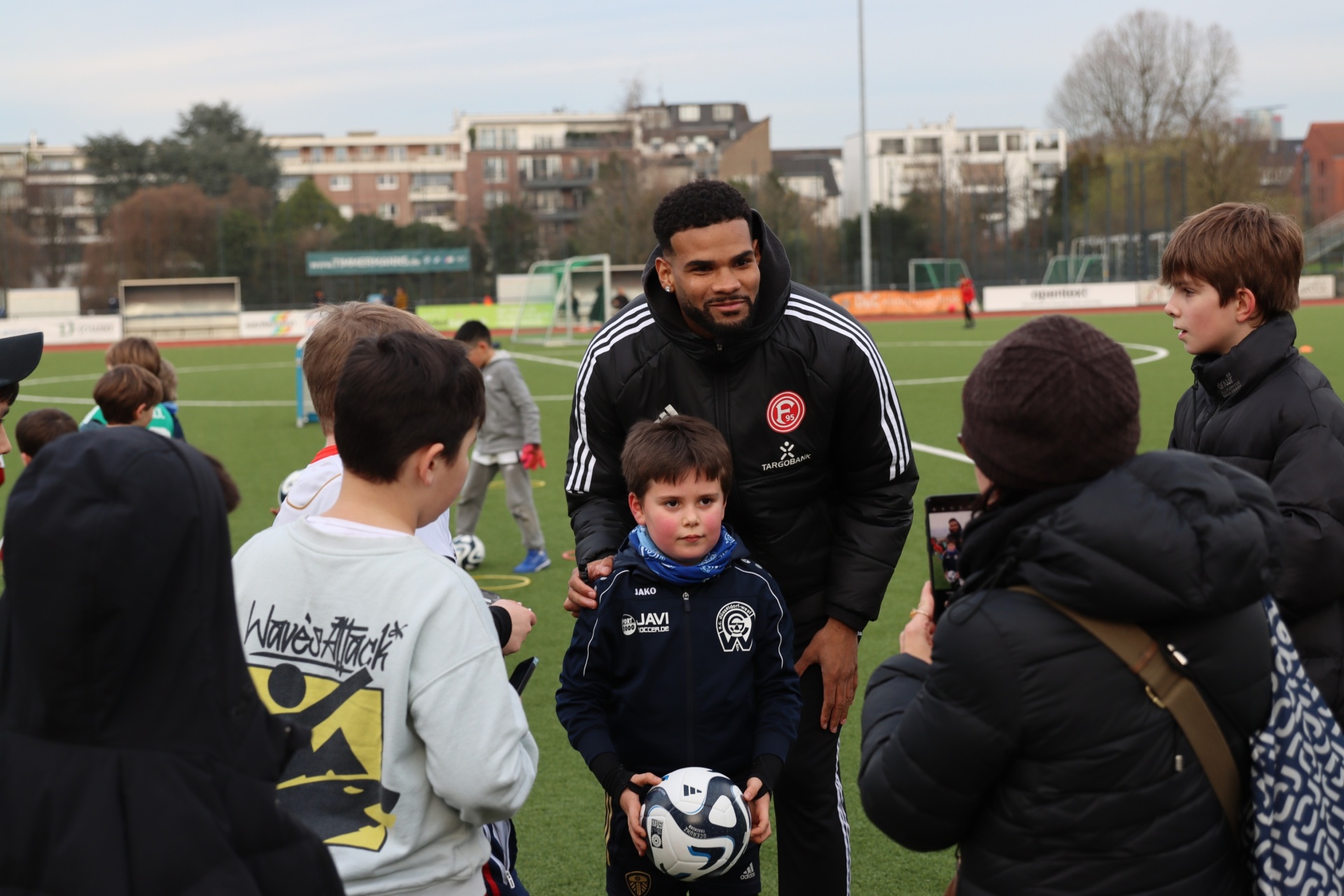 [{"clang_id":1,"value":"Fußballspieler umringt von Kindern auf einem Fußballplatz. Er unterhält sich mit einem Jungen, der einen Fußball hält. Im Hintergrund weitere Kinder und Gebäude."}]