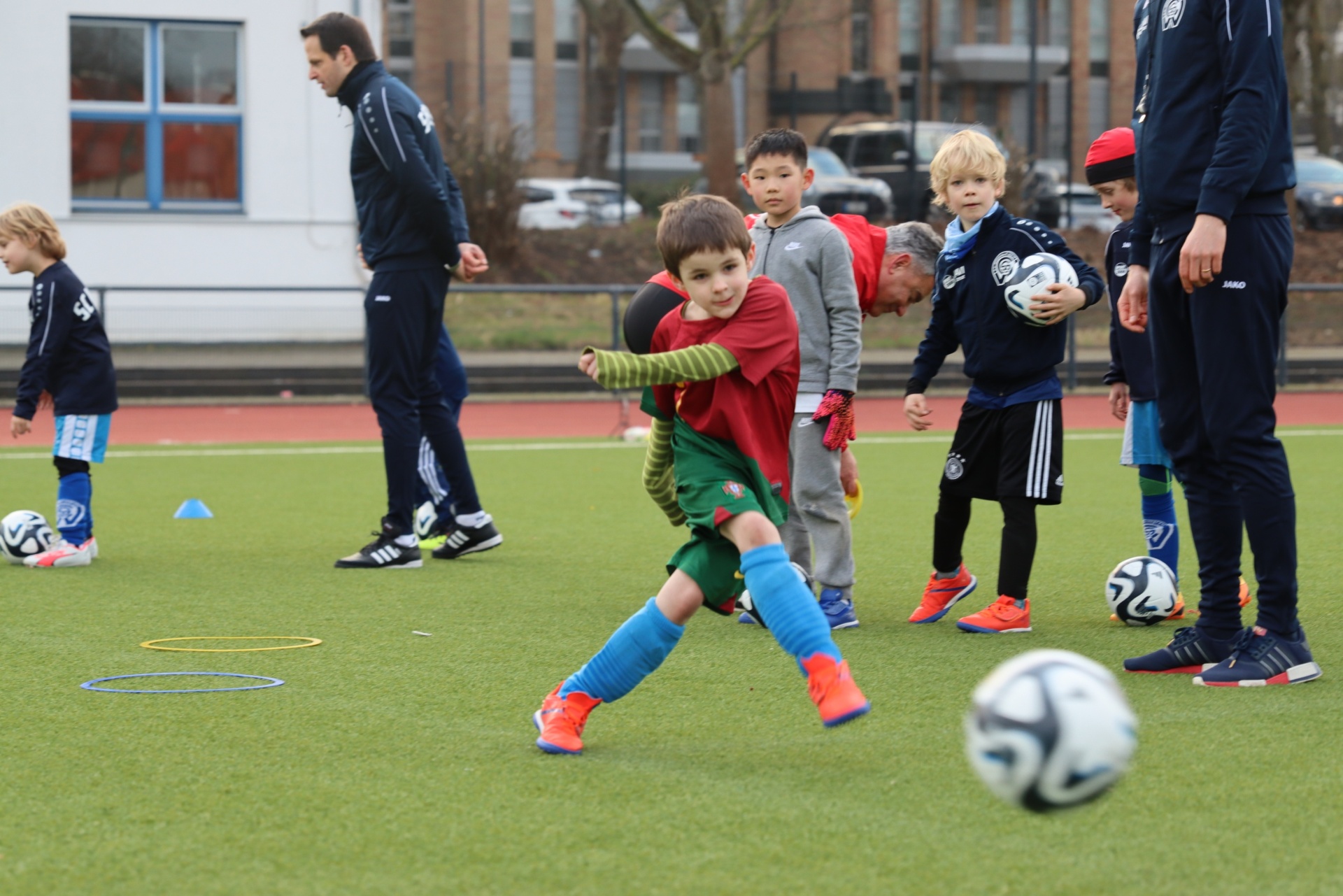 [{"clang_id":1,"value":"Fußballtraining mit Kindern: Ein Junge schießt auf ein Fußballfeld, andere Kinder stehen um ihn herum, zwei Trainer beobachten. Gebäude im Hintergrund."}]