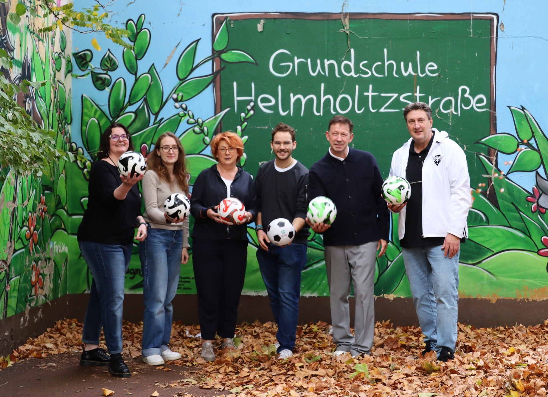 [{"clang_id":1,"value":"Gruppe von sechs Personen vor einer Schulwand mit der Aufschrift \"Grundschule Helmholtzstraße\". Alle halten Fußbälle. Herbstliche Blätter am Boden."}]