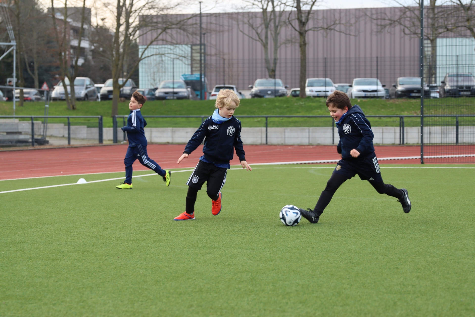 [{"clang_id":1,"value":"Drei Kinder spielen Fußball auf einem grünen Kunstrasenplatz. Sie tragen blaue Trainingsanzüge. Im Hintergrund eine Sportanlage und parkende Autos."}]