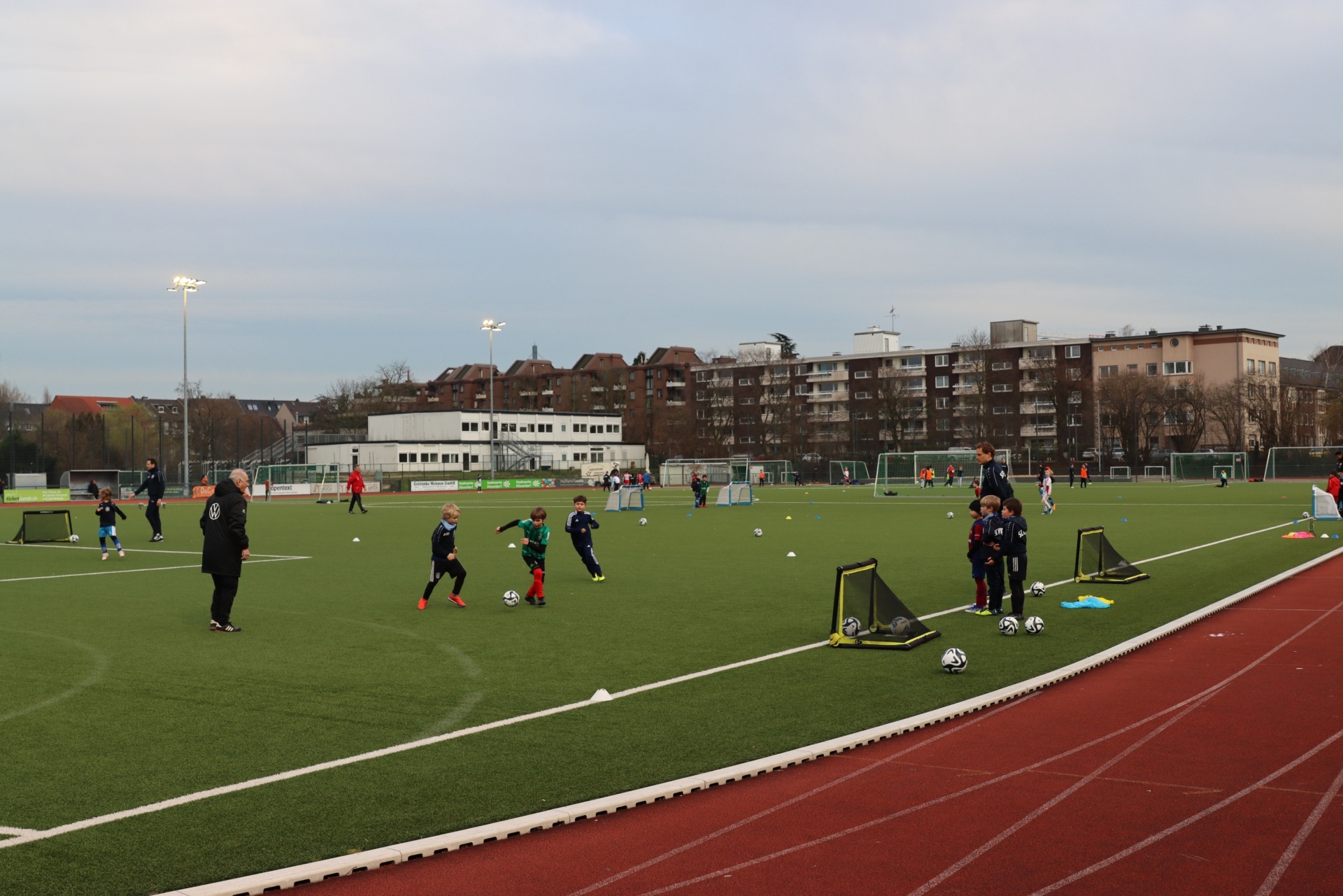 [{"clang_id":1,"value":"Fußballfeld mit trainierenden Kindern. Im Hintergrund Wohngebäude und Lichtmasten. Roter Laufbahnrand."}]