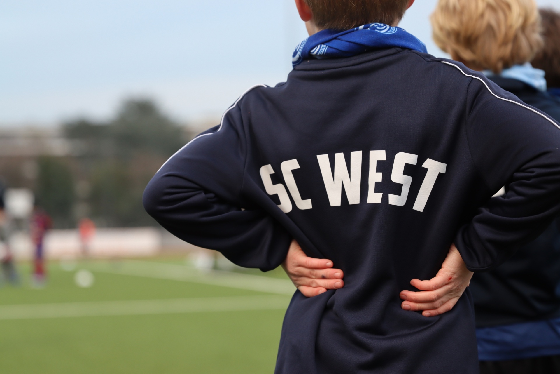 [{"clang_id":1,"value":"Rückansicht eines jungen Mannes mit blauem Trainingsanzug und Aufschrift \"SC WEST\", der mit den Händen in den Hüften auf einem Fußballfeld steht."}]