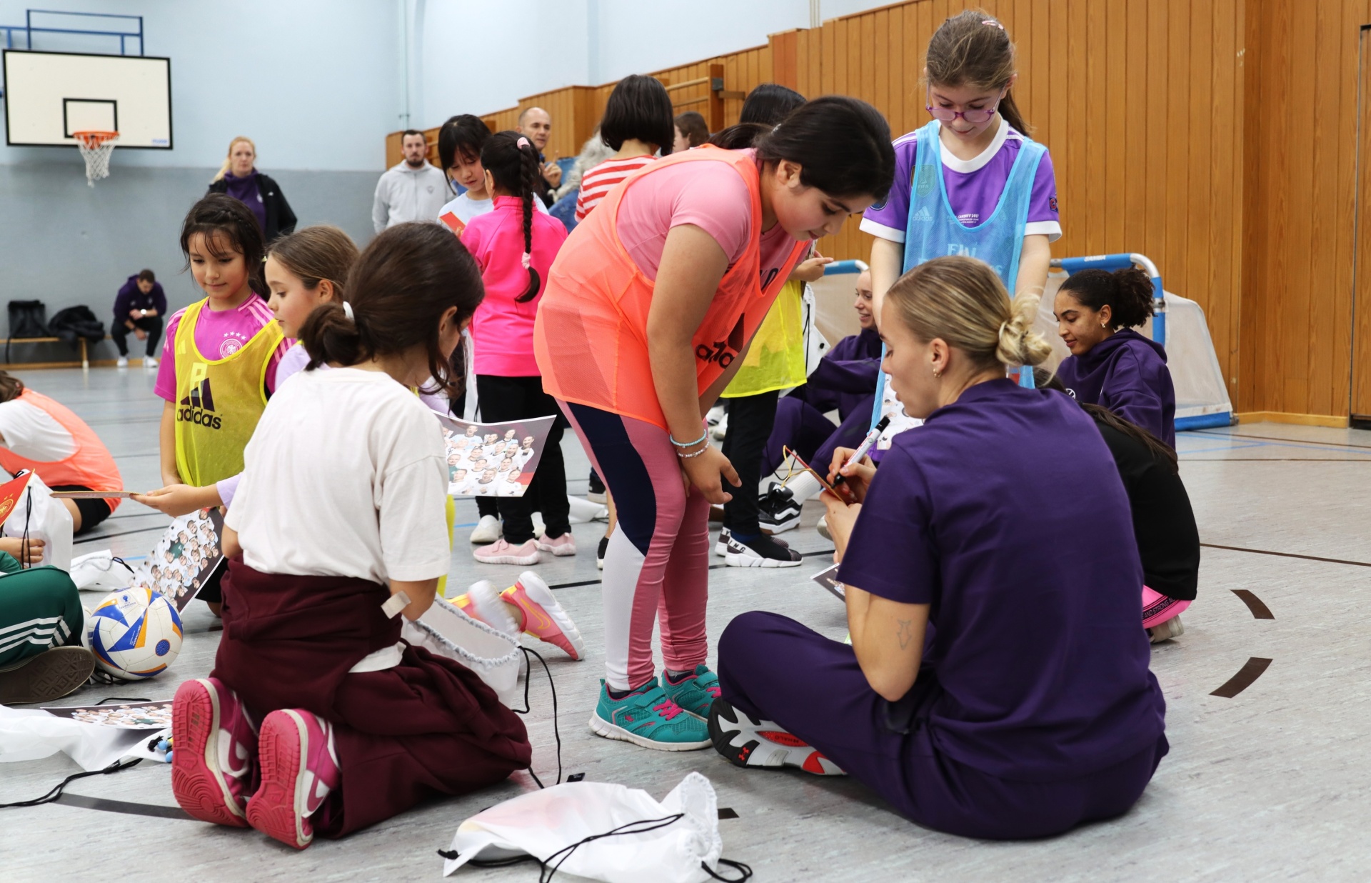 [{"clang_id":1,"value":"Sporthalle. Gruppe Mädchen und Frauen knien auf dem Boden und prüfen Sportschuhe. Einige tragen Trainingskleidung, andere violette Oberteile. Im Hintergrund weitere Personen und ein Basketballkorb."}]