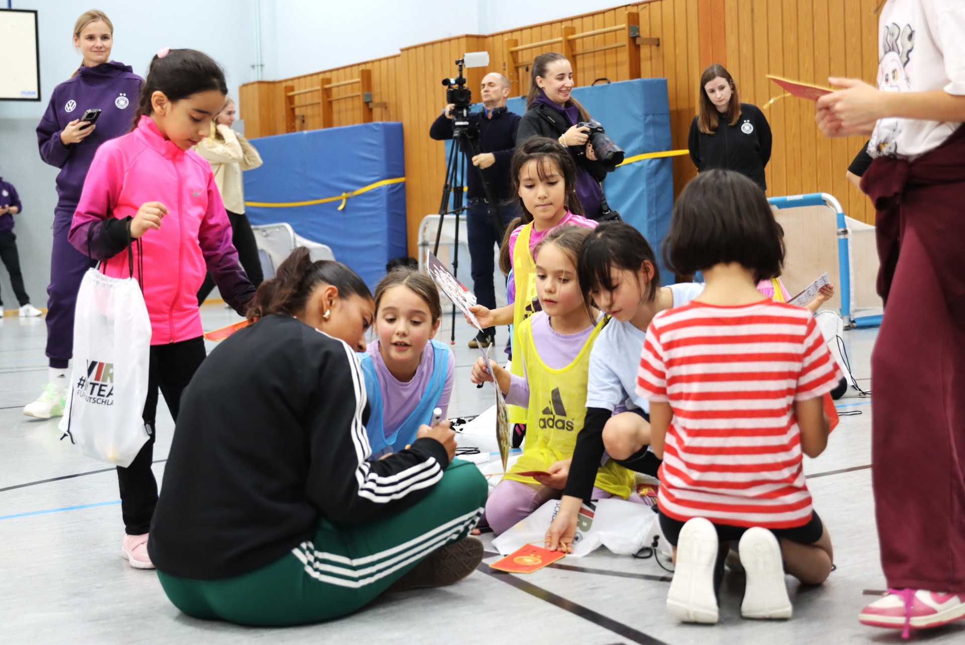 [{"clang_id":1,"value":"Frau kniet vor Kindern in Sporthalle, unterschreibt Karten. Im Hintergrund weitere Personen und blaue Matten."}]