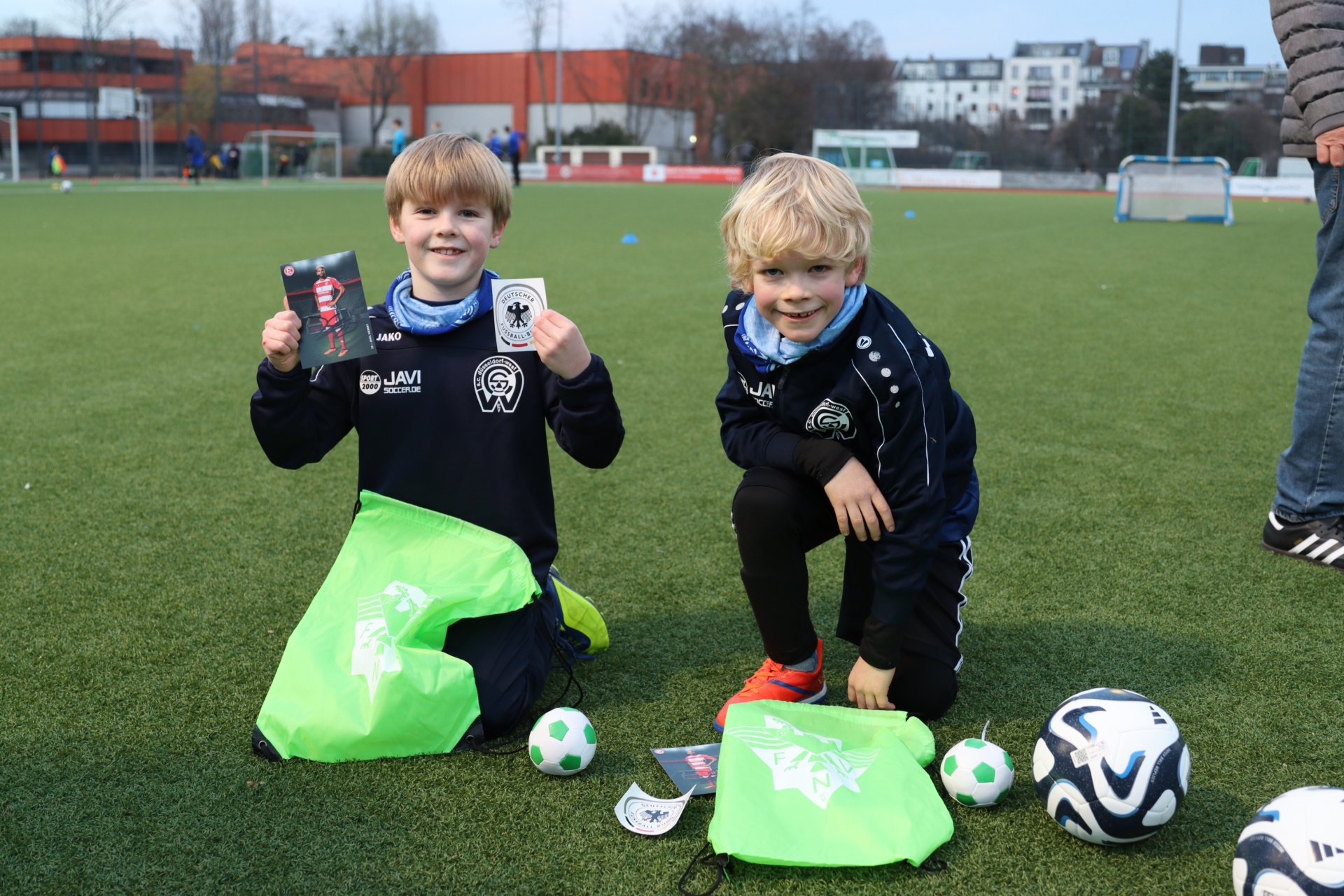 [{"clang_id":1,"value":"Zwei Jungen knien auf einem Fußballfeld, öffnen Geschenktüten und halten kleine Fußball-Souvenirs. Im Hintergrund ein Fußballfeld und Gebäude."}]