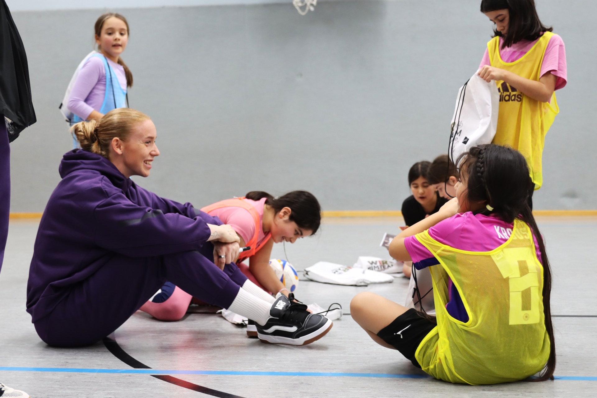 [{"clang_id":1,"value":"Frau sitzt auf dem Boden und hilft einem Mädchen, Schuhe zuzubinden. Mehrere Kinder in Sportkleidung sitzen oder stehen im Hintergrund in einer Sporthalle."}]