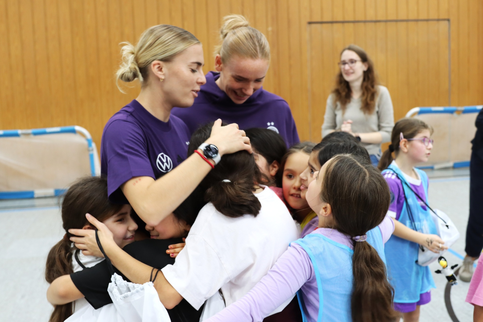 [{"clang_id":1,"value":"Zwei Fußballspielerinnen in lila Trainingsanzügen umarmen eine Gruppe junger Mädchen in einem Hallensportraum. Im Hintergrund stehen weitere Personen."}]