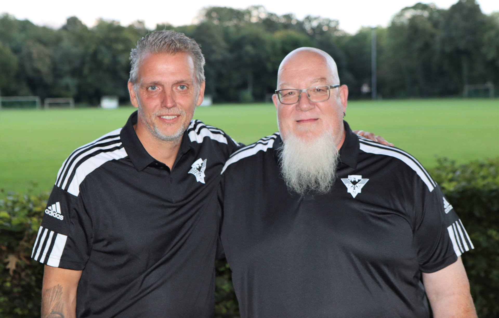 [{"clang_id":1,"value":"Zwei Männer mit Polo-Shirts vor einem Fußballfeld. Links ein Mann mit kurz grauem Haar, rechts ein Mann mit Bart und Glatze. Beide lächeln."}]