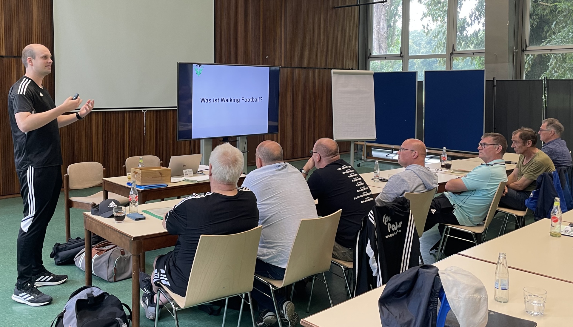 [{"clang_id":1,"value":"Mann steht vor Gruppe sitzender Männer an Tischen. Im Hintergrund eine Projektion mit der Frage \"Was ist Walking Football?\". Innenraum mit Fenstern und Bäumen im Hintergrund."}]