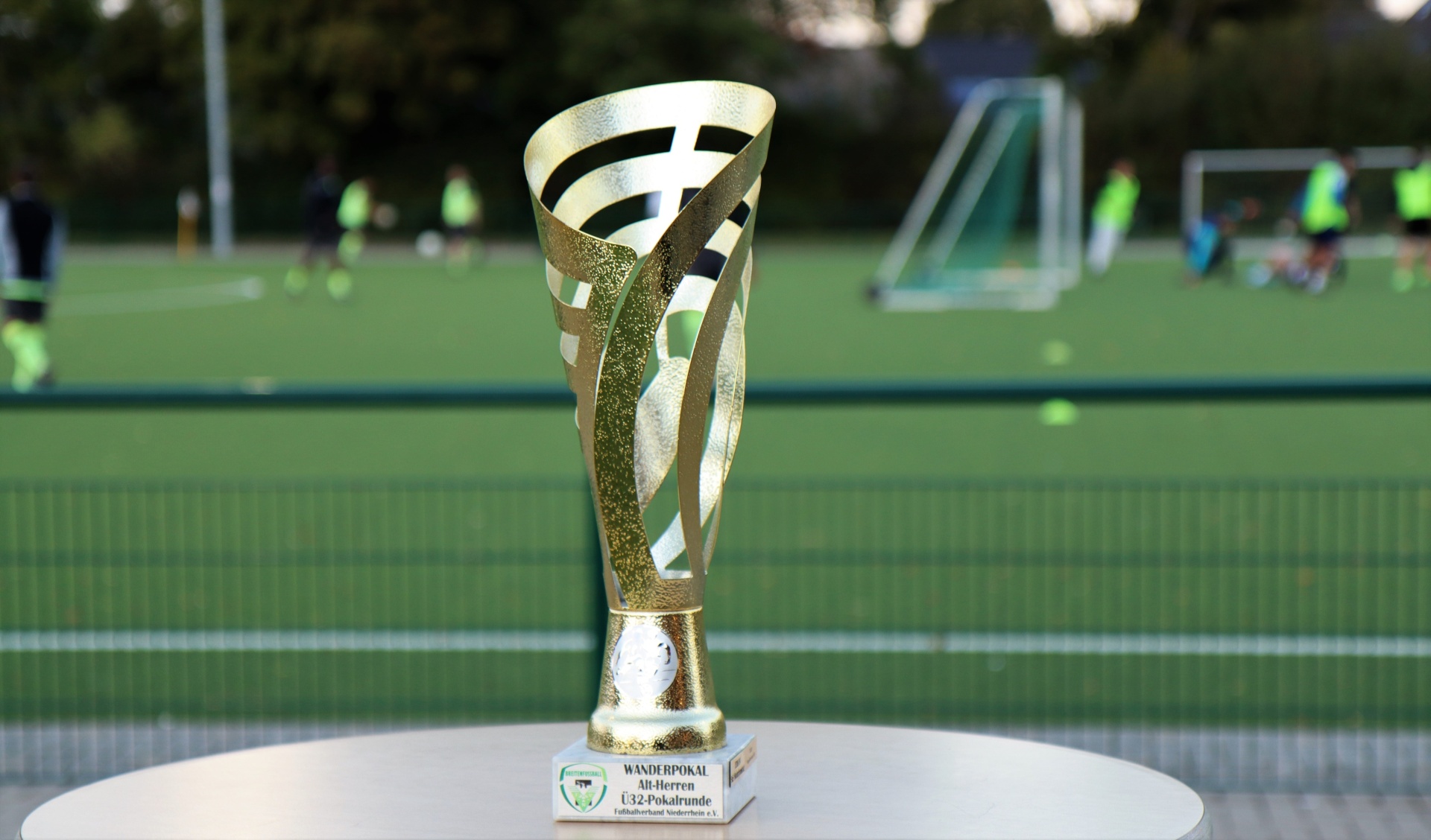 [{"clang_id":1,"value":"Goldener Wanderpokal auf einem Podest vor einem Hockeyfeld mit Spielern und Toren im Hintergrund. Beschriftung am Podest."}]