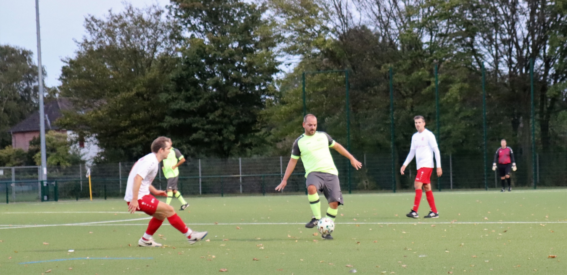[{"clang_id":1,"value":"Fußballspiel im Freien: Drei Spieler mit Ball auf grünem Kunstrasenfeld, umgeben von Zuschauern und Bäumen."}]